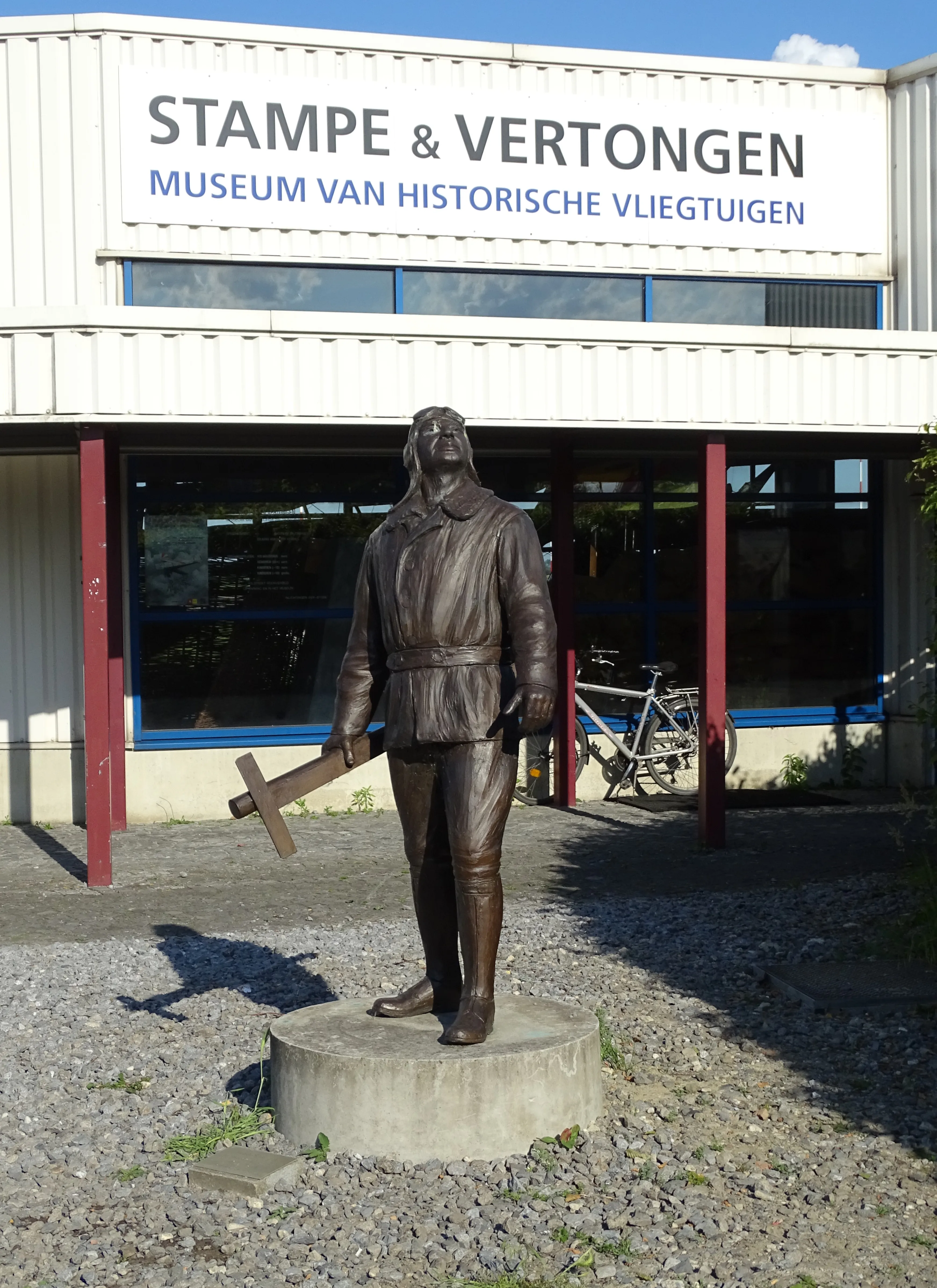 Estatua de bronce de un aviador sobre una base de hormigón frente al edificio del Stampe & Vertongen Museum, con un gran cartel que dice 'STAMPE & VERTONGEN MUSEUM VAN HISTORISCHE VLIEGTUIGEN' sobre la entrada.