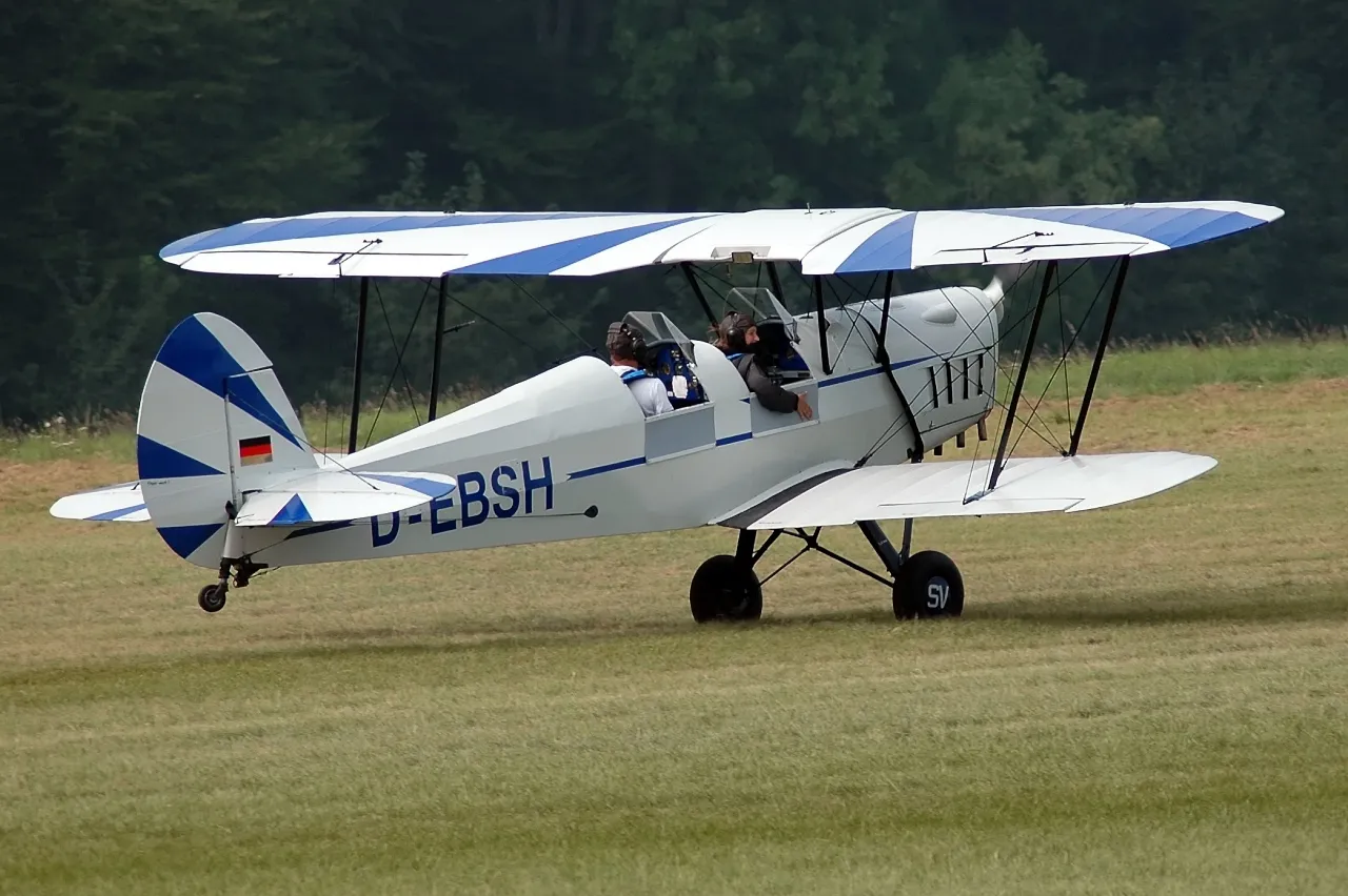 Biplano Stampe-Vertongen SV-4 blanco y azul con registro D-EBSH rodando por un aeródromo de hierba con dos ocupantes visibles en la cabina abierta