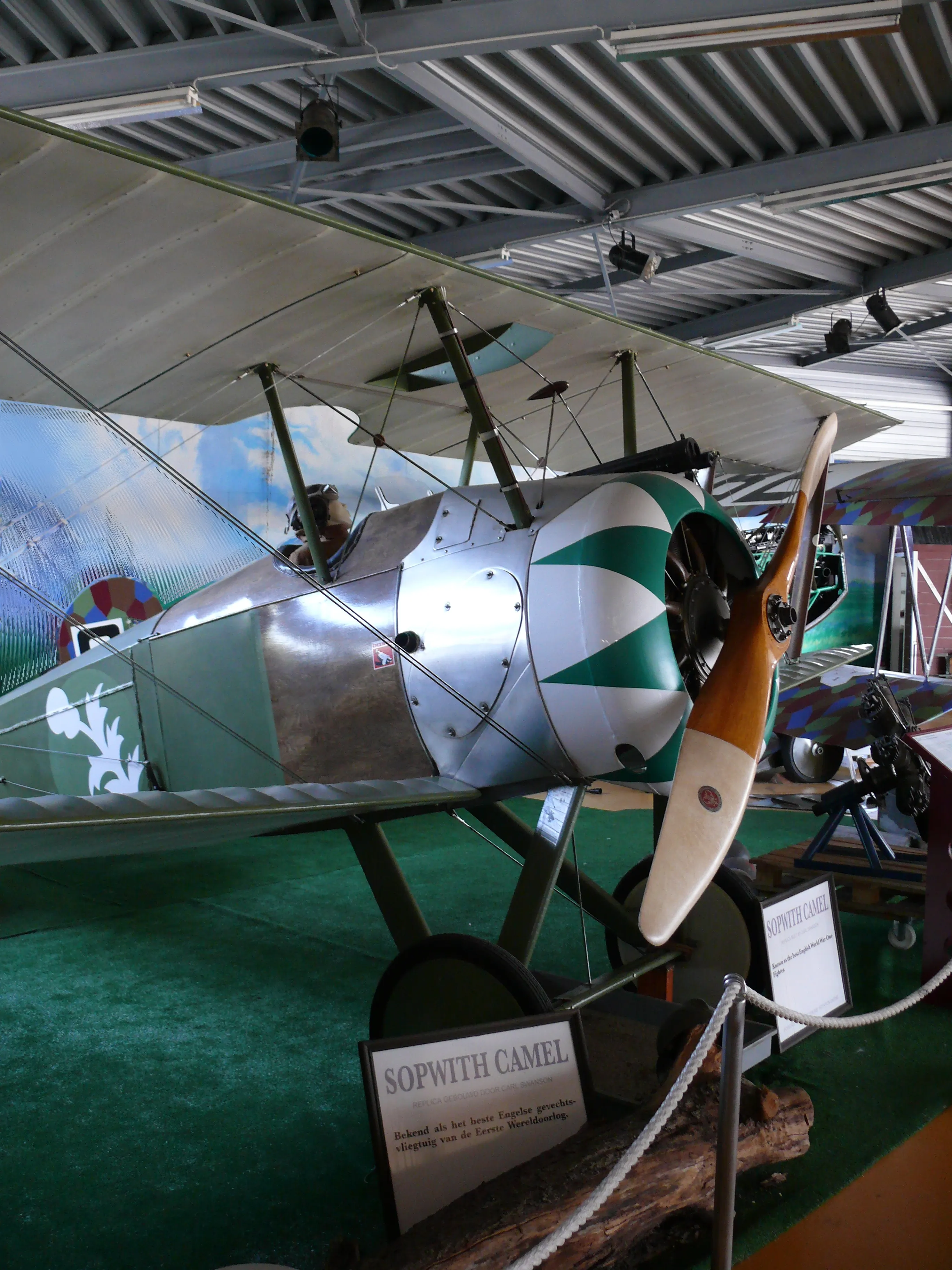 Un biplano Sopwith Camel con marcas verdes y blancas en el morro y una hélice de madera expuesto en un museo, con un cartel que dice SOPWITH CAMEL delante.