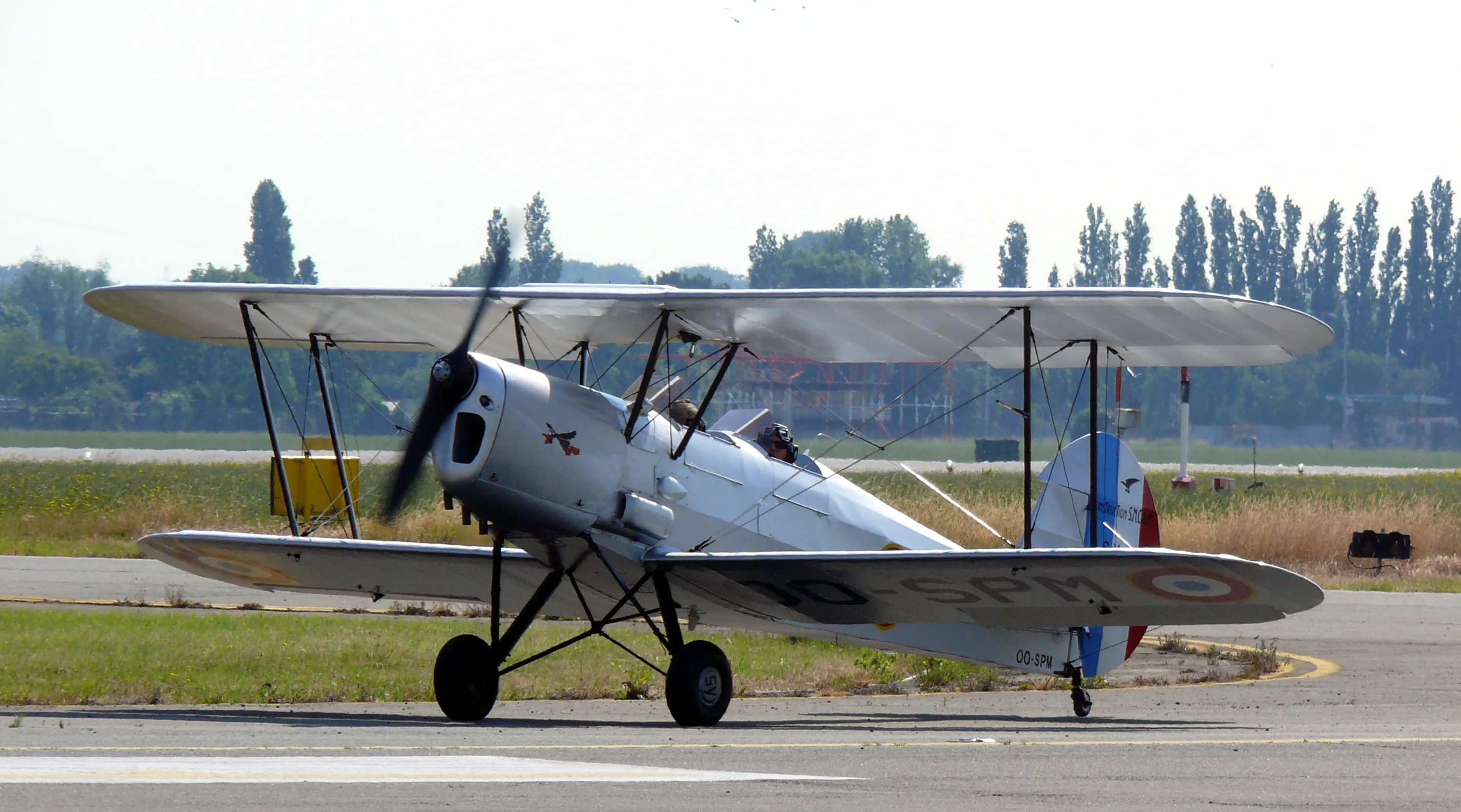 Biplano Silver Stampe & Vertongen SV.4C rodando por una pista con una hélice giratoria borrosa y una hilera de árboles al fondo.