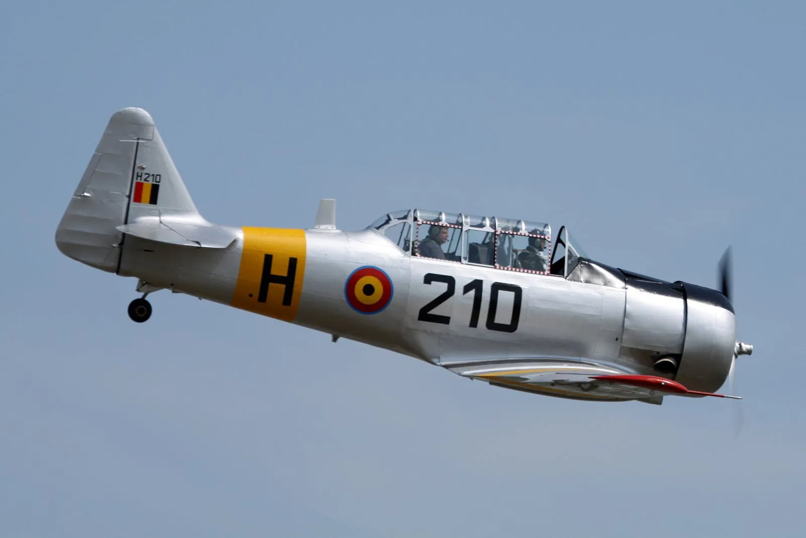 Un avión vintage plateado Stampe et Vertongen con marcas H 210, una escarapela y la bandera belga en la cola, volando contra un cielo azul despejado.