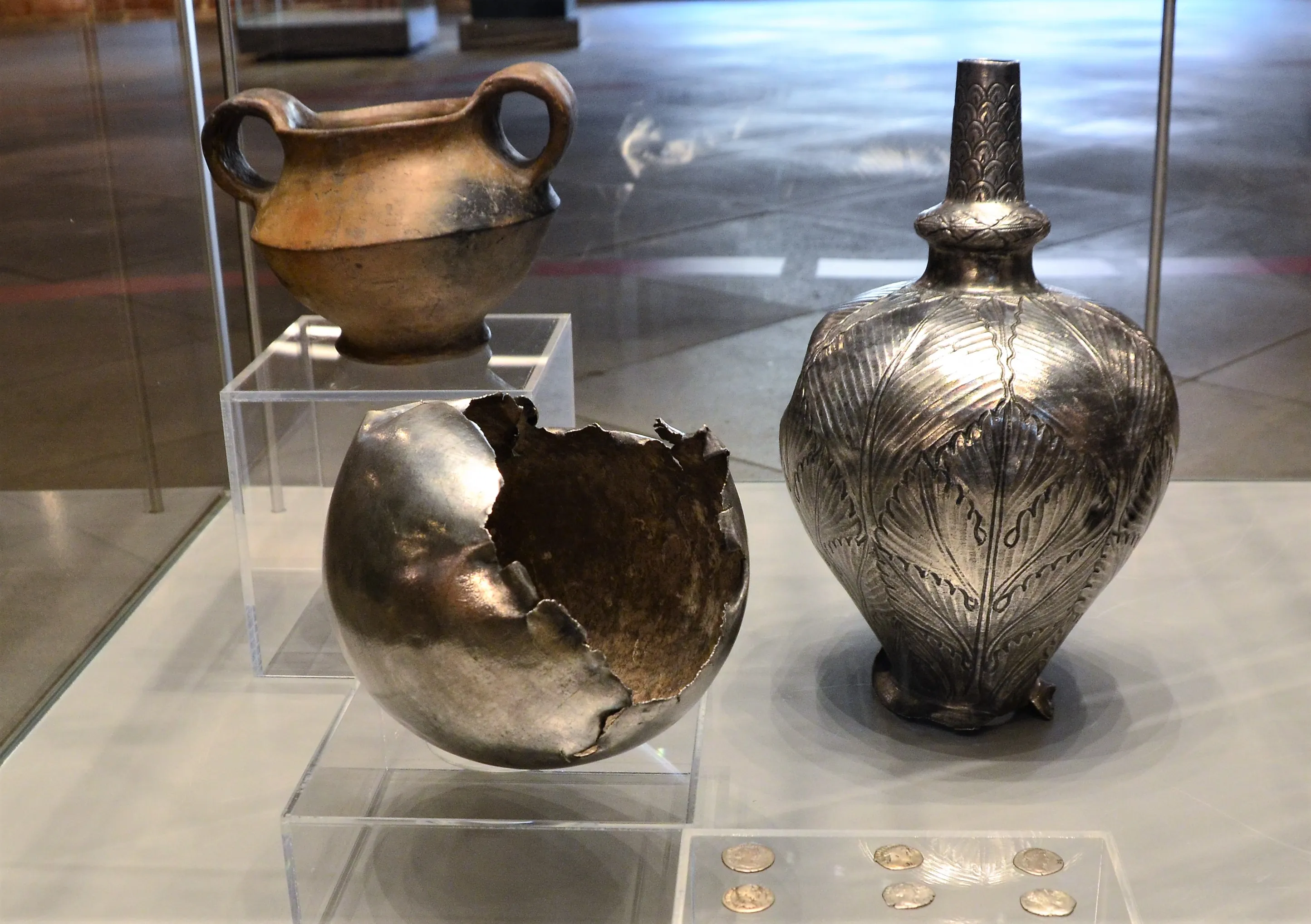 three ancient artifacts in a glass display case including a broken metallic bowl, a tall decorated vase, and a two-handled pot