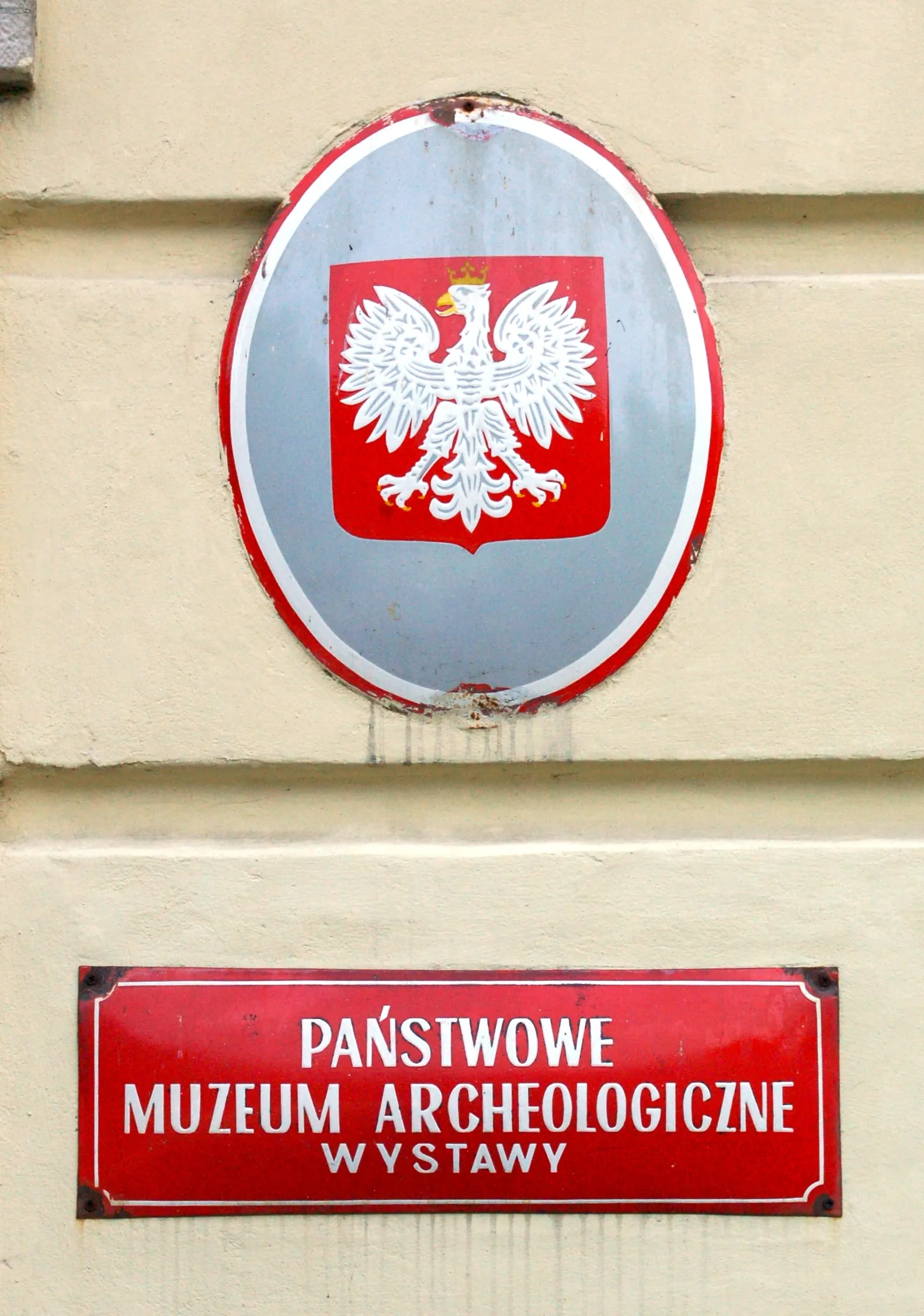 Red sign with white text reading 'PAŃSTWOWE MUZEUM ARCHEOLOGICZNE WYSTAWY' below a circular emblem with a white eagle on a red shield.