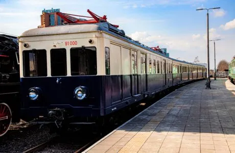 Niebiesko-kremowy pociąg elektryczny z numerem 91 000 z przodu, zaparkowany na torach na peronie stacji.