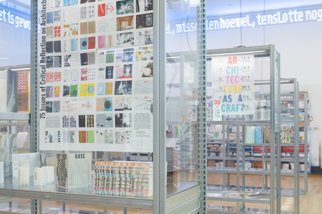 Museuminterieur met glazen vitrines met boeken en tentoonstellingsposters, inclusief 'Architecture as a Craft' bewegwijzering