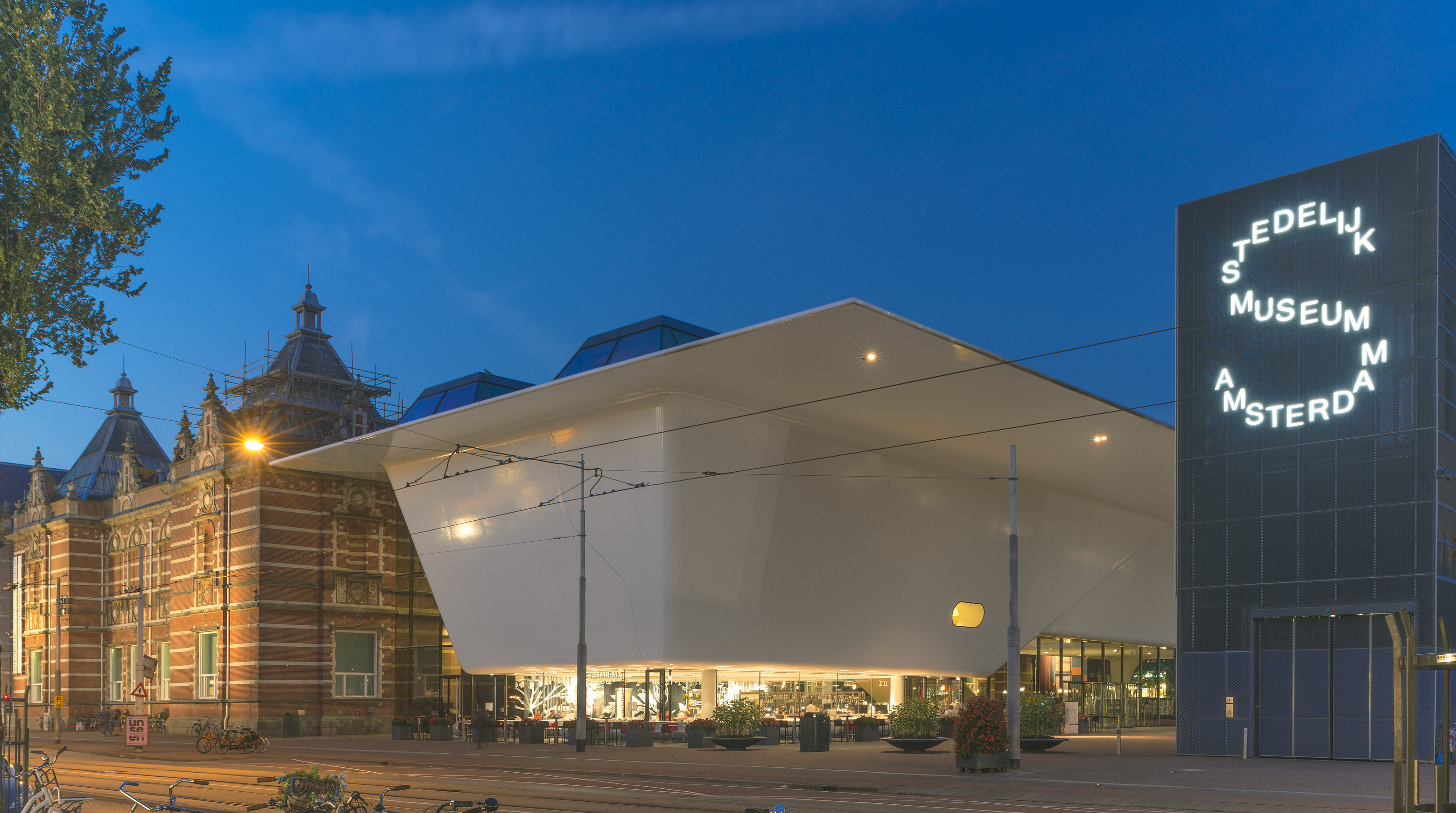 Stedelijk Museum Amsterdam exterieur bij schemering, met moderne witte uitbreiding, historisch bakstenen gebouw en verlicht cirkelvormig uithangbord.