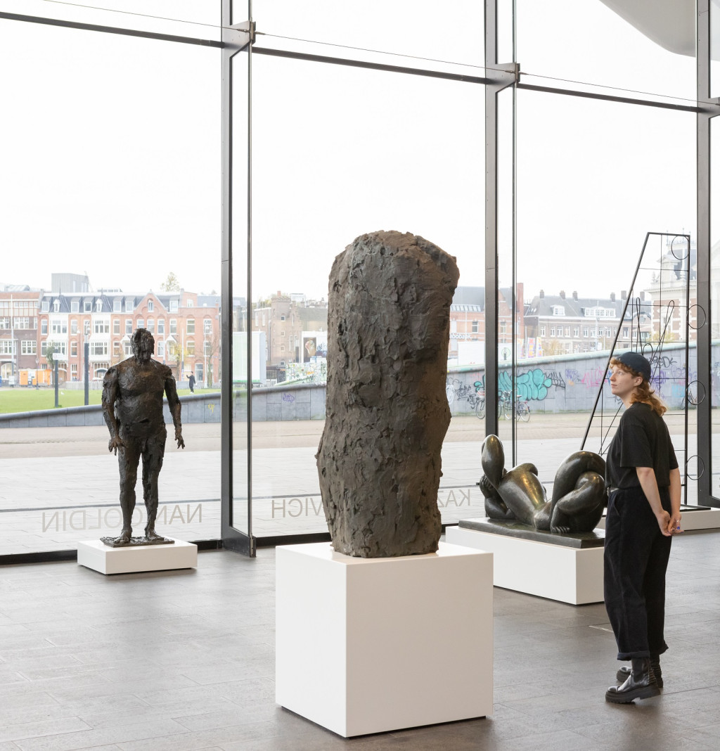 Moderne kunstgalerie met kamerhoge ramen die uitzicht op de stad bieden, met drie sculpturen op witte sokkels en een bezoeker die de tentoonstellingen bekijkt.