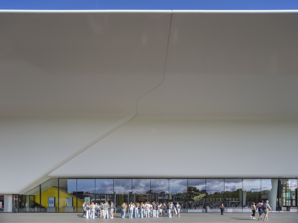 Modern white building facade with curved roof and large glass entrance of Stedelijk Museum Amsterdam, people gathered outside on a sunny day