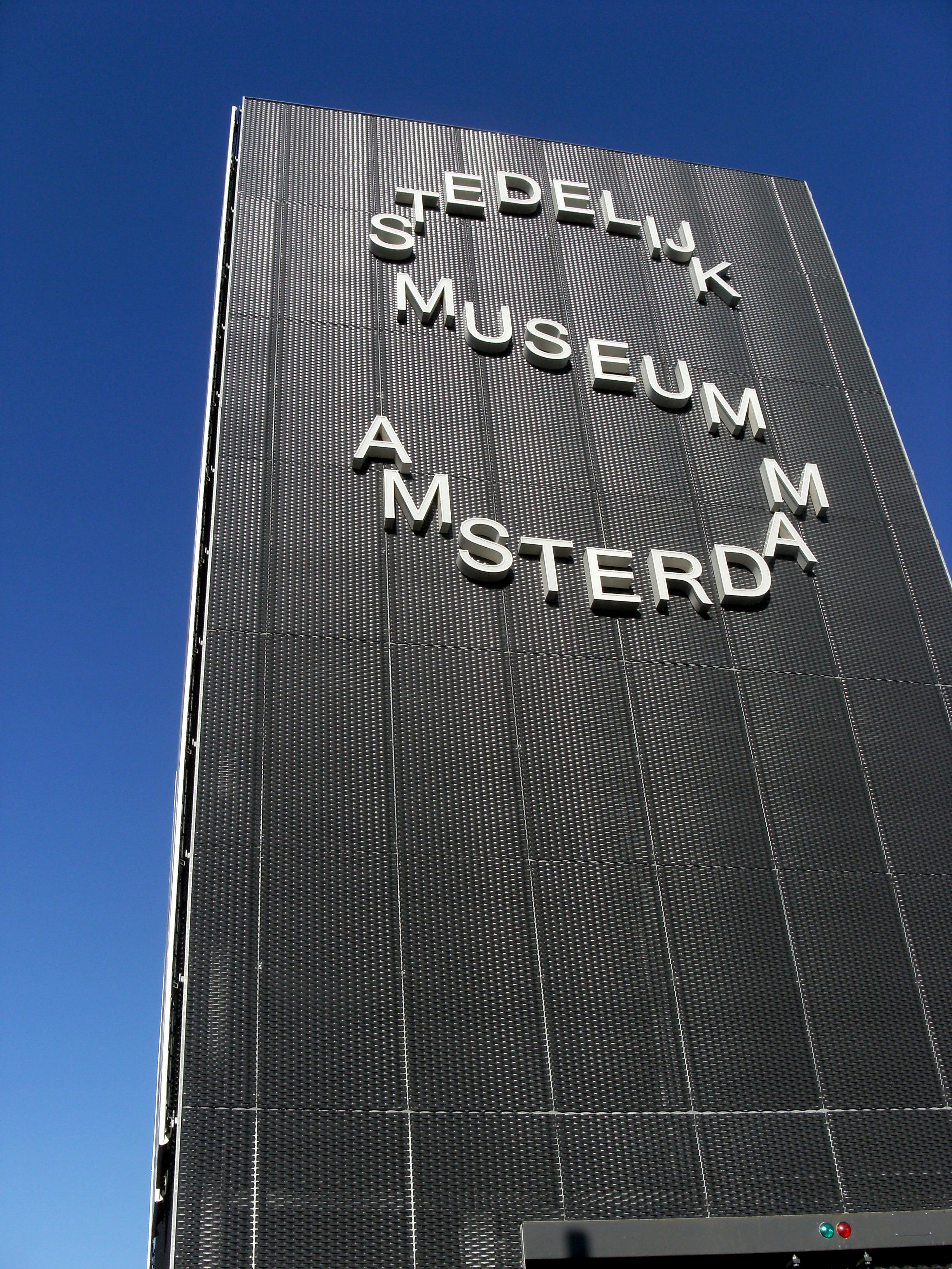 Het Stedelijk Museum Amsterdam-bord op een donkere, getextureerde gevel onder een blauwe lucht