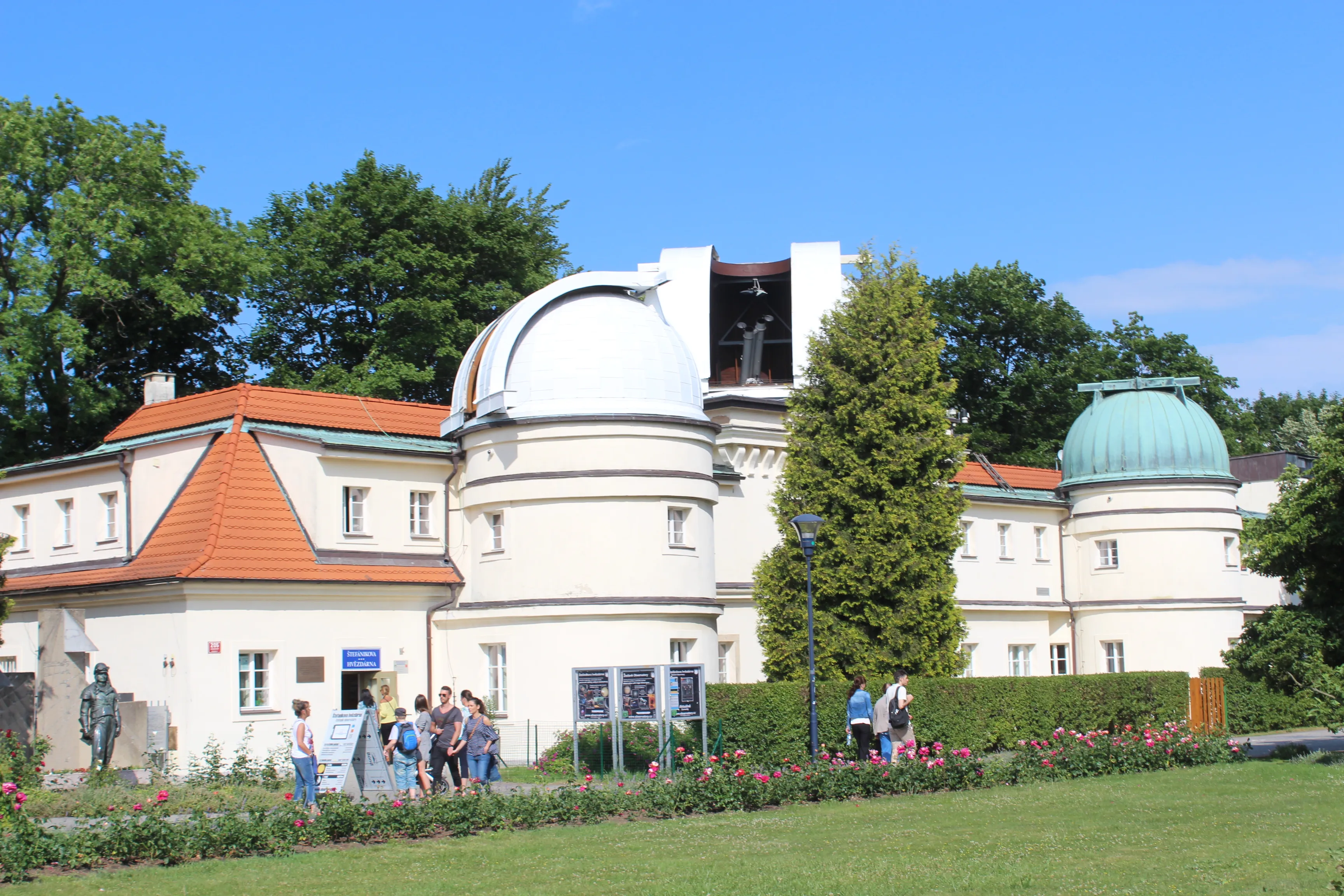 Štefánik Observatory with white domes and a red-roofed building, surrounded by green trees and grass, with people walking nearby.