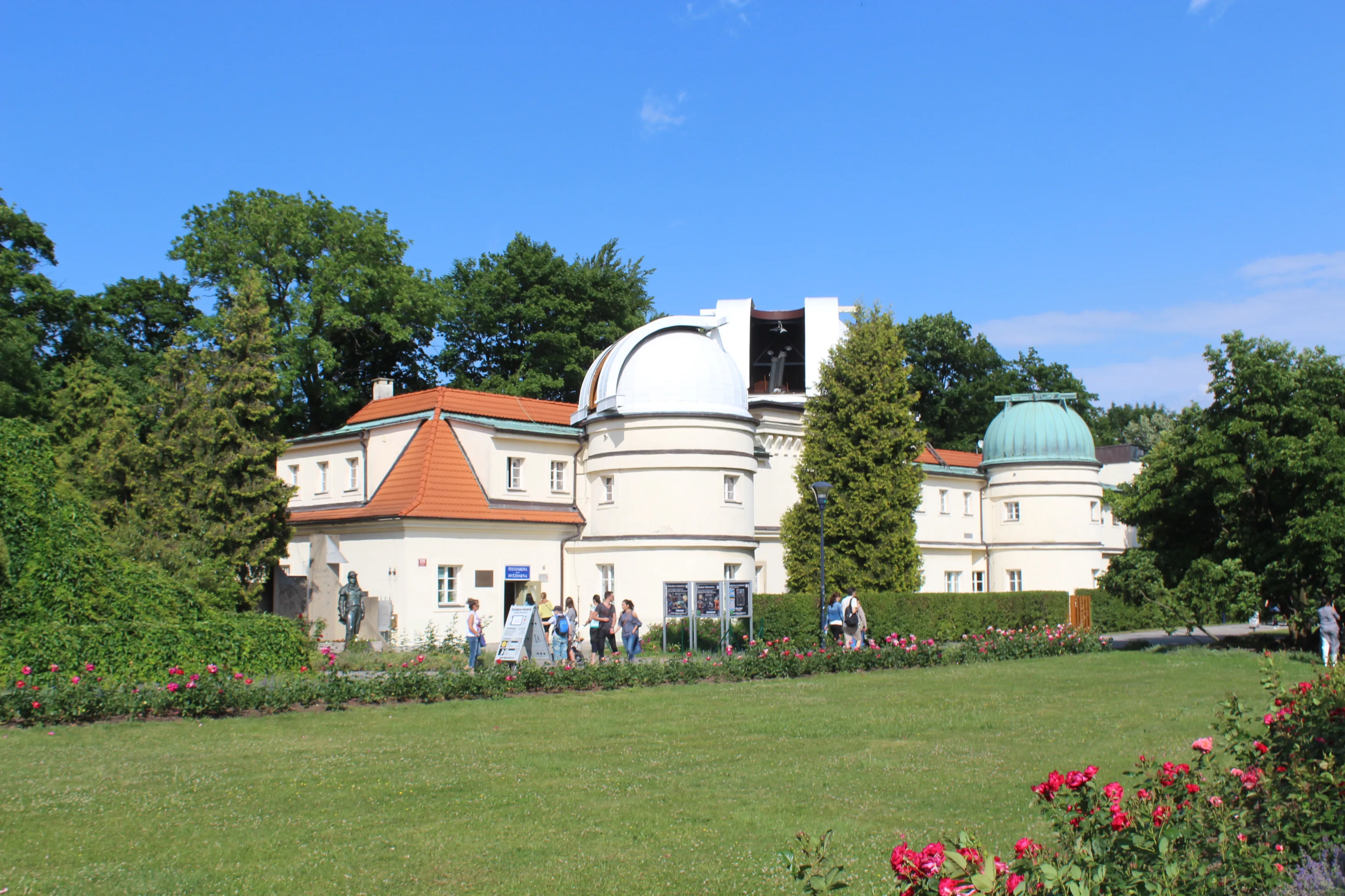 Observatorio Štefánik con edificios de telescopios de cúpula blanca y una estructura con techo rojo, rodeado de césped verde y árboles bajo un cielo azul