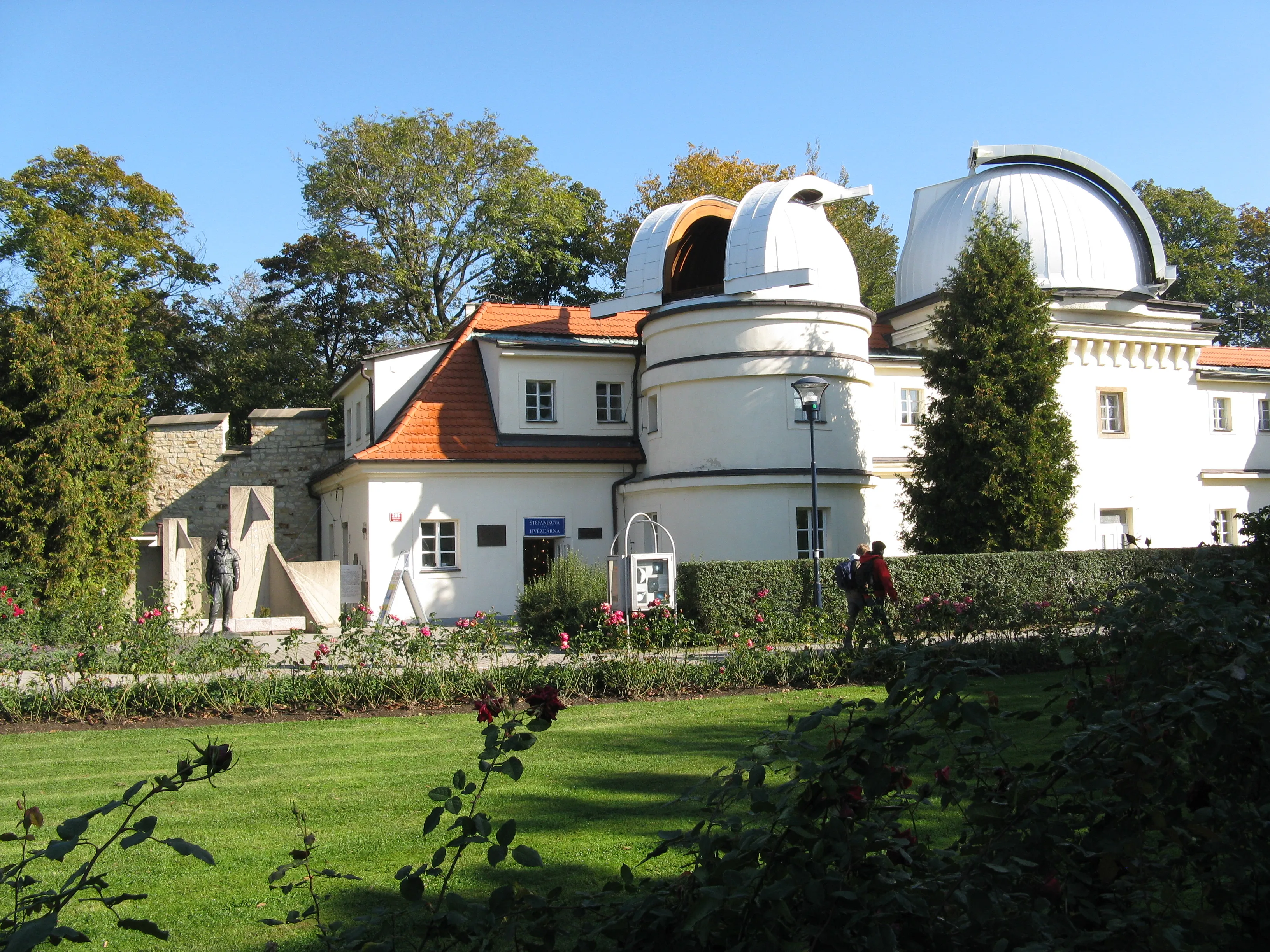 Edificio del observatorio blanco con dos estructuras de telescopio abovedadas, rodeado de hierba verde y árboles bajo un cielo azul