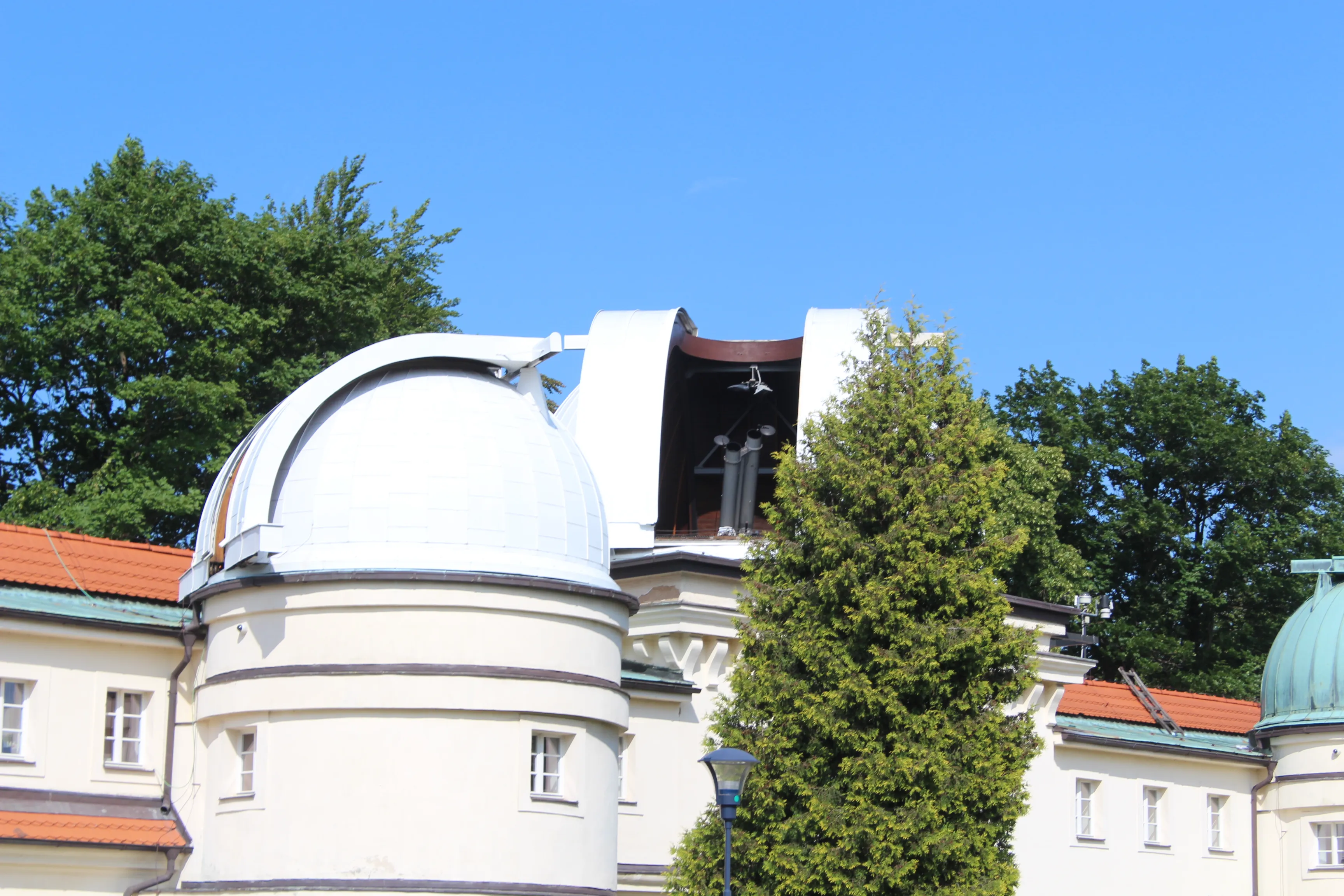 Cúpula de observatorio blanco con tapa abierta, rodeada de árboles y un cielo azul claro
