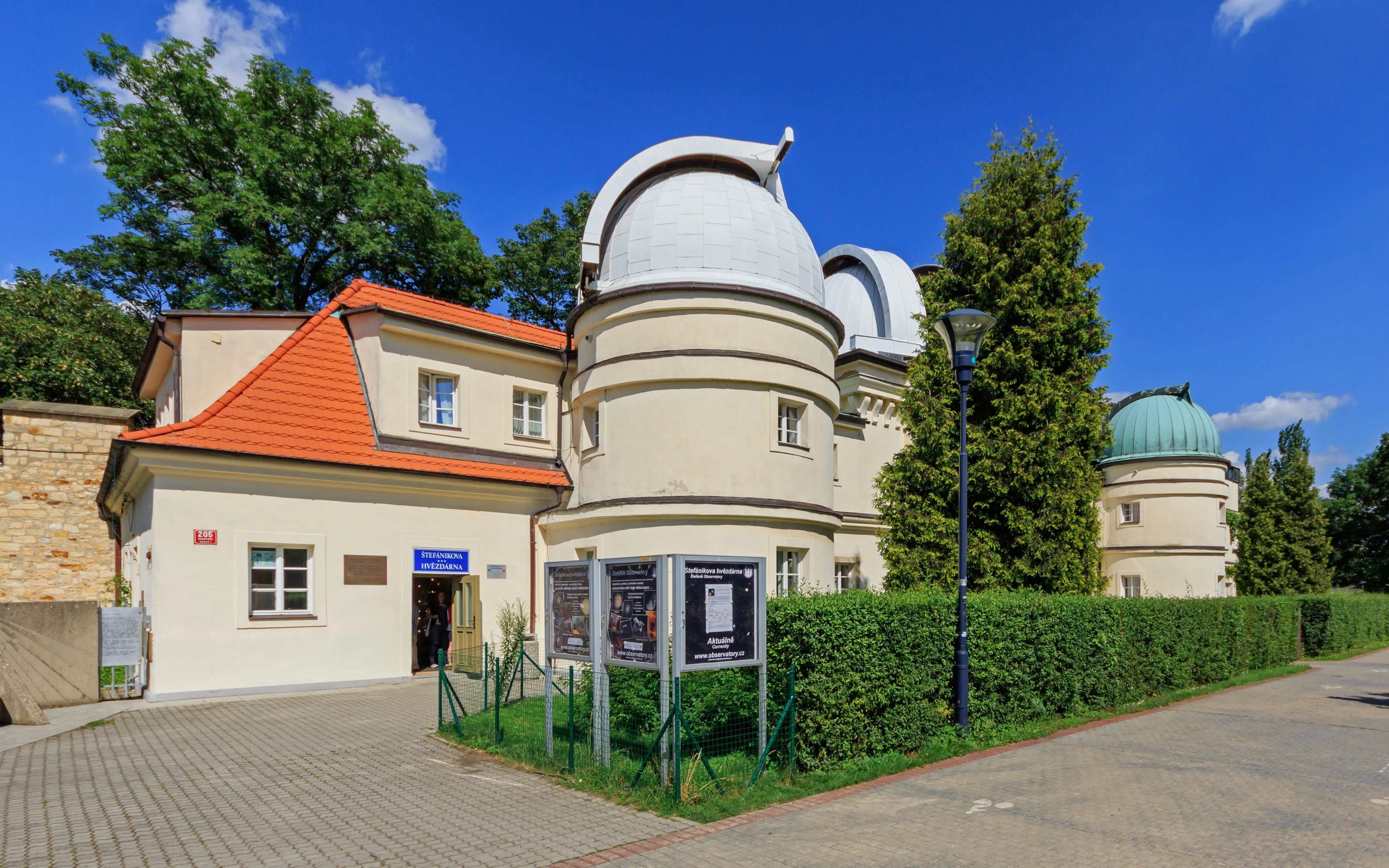 Observatorio Štefánik con edificios de observatorio de cúpula blanca y una estructura con techo rojo rodeada de árboles verdes y setos bajo un cielo azul
