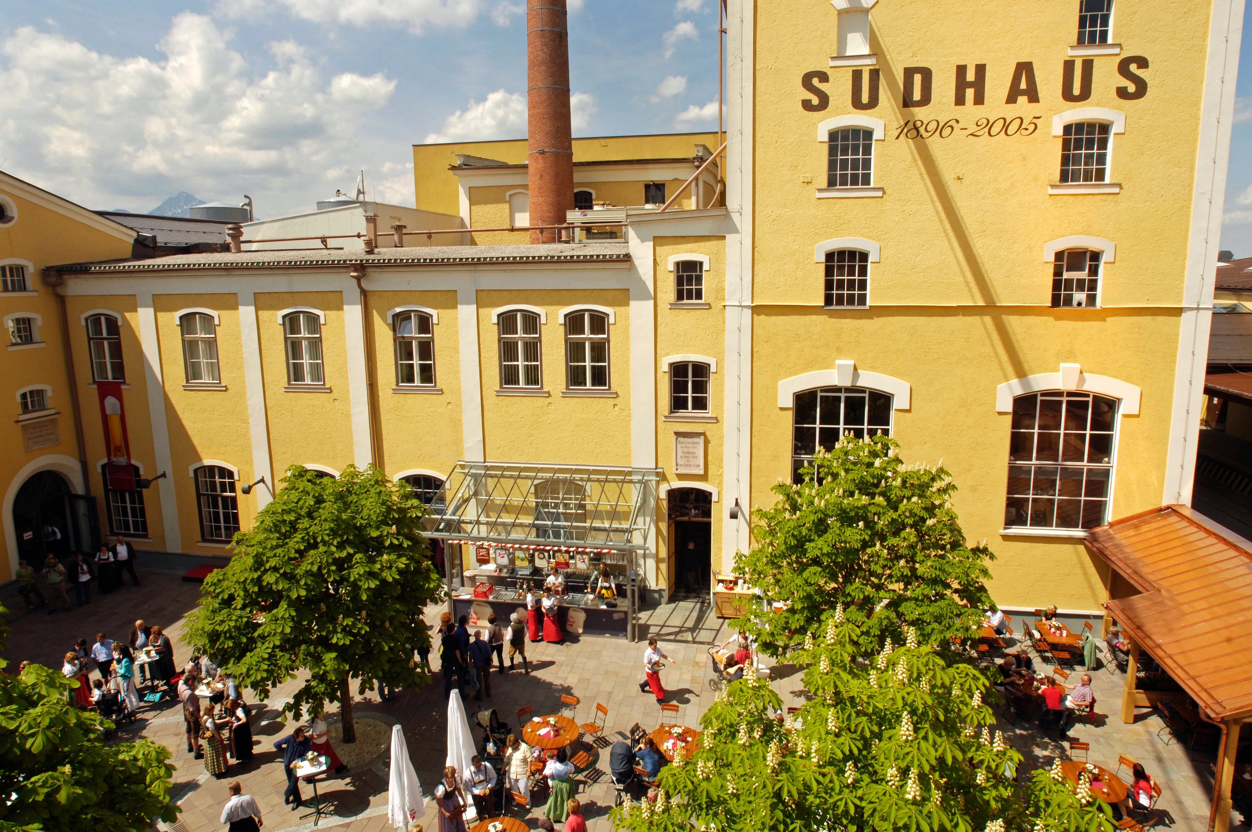 Vista aérea de los edificios amarillos de la cervecería Stiegl con el letrero "SUDHAUS 1890-2005", patio con árboles y visitantes.