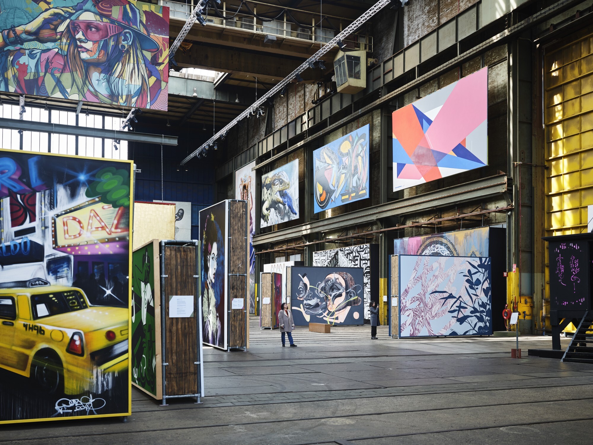 Grote industriële ruimte met levendige street art-muurschilderingen, een geeltaxi-sculptuur en een bezoeker die tussen tentoonstellingen wandelt