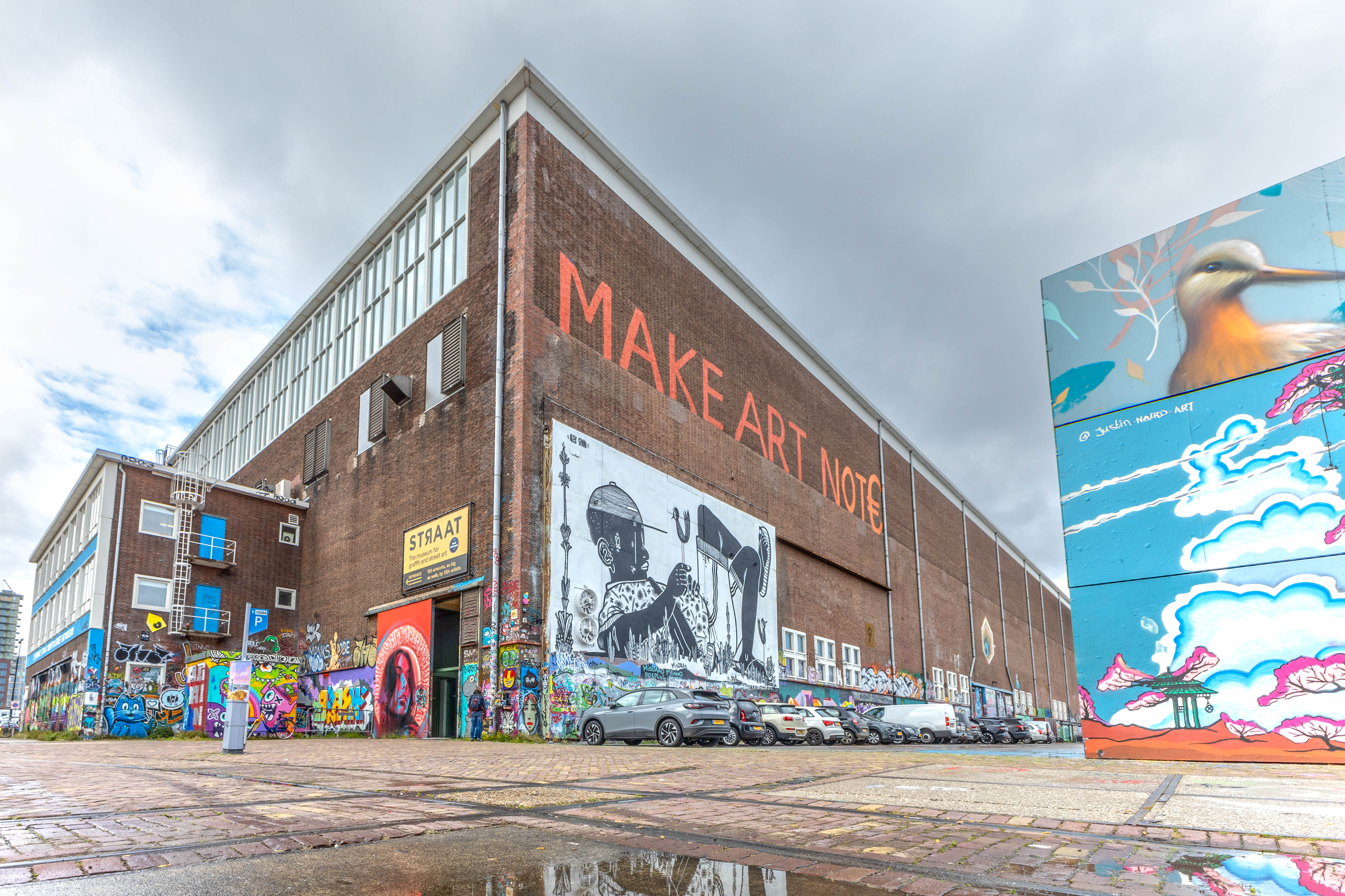Bakstenen industrieel gebouw met de tekst 'MAKE ART NOT' en kleurrijke muurschilderingen, waaronder een groot schilderij van een vogel, op de buitenmuren onder een bewolkte hemel