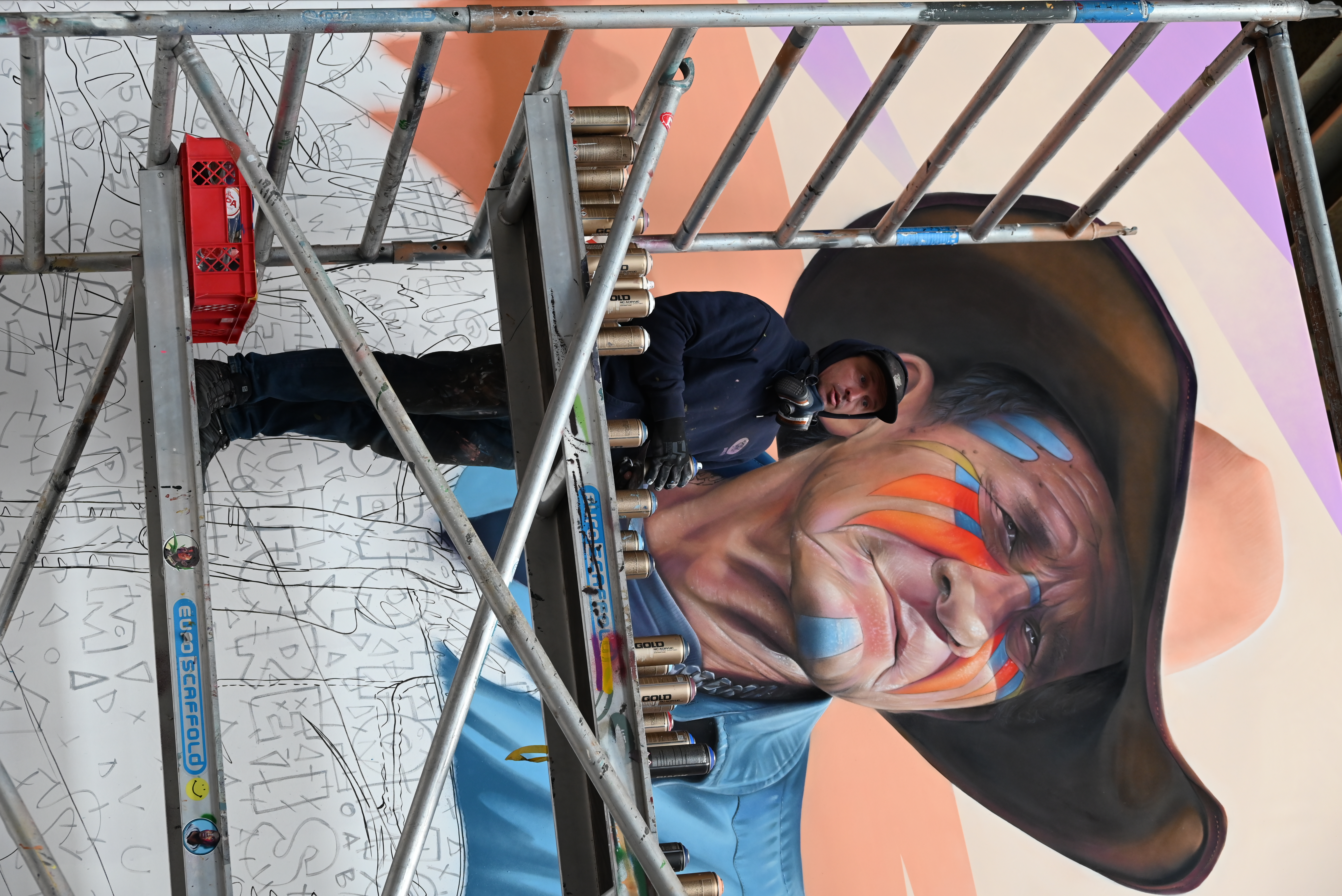 Artysta na rusztowaniu tworzy duży mural przedstawiający osobę z kolorowym makijażem twarzy i kapeluszem z szerokim rondem w muzeum STRAAT