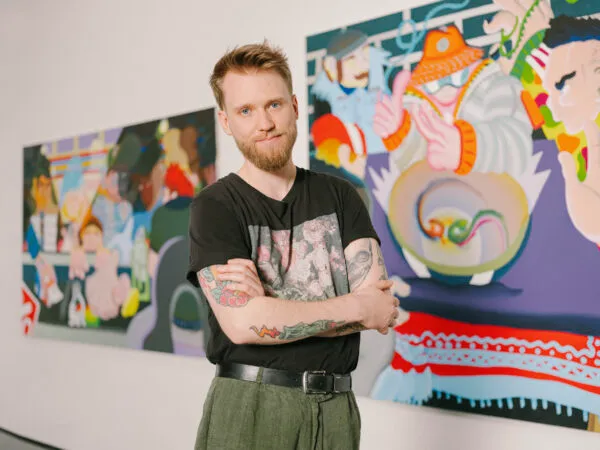 Mężczyzna z tatuażami stoi z założonymi rękami przed kolorowymi współczesnymi obrazami w STRABAG Kunstforum.