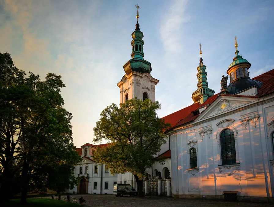 Biały budynek w stylu barokowym z zielonymi iglicami i czerwonym dachem pod błękitnym niebem z drzewami na pierwszym planie