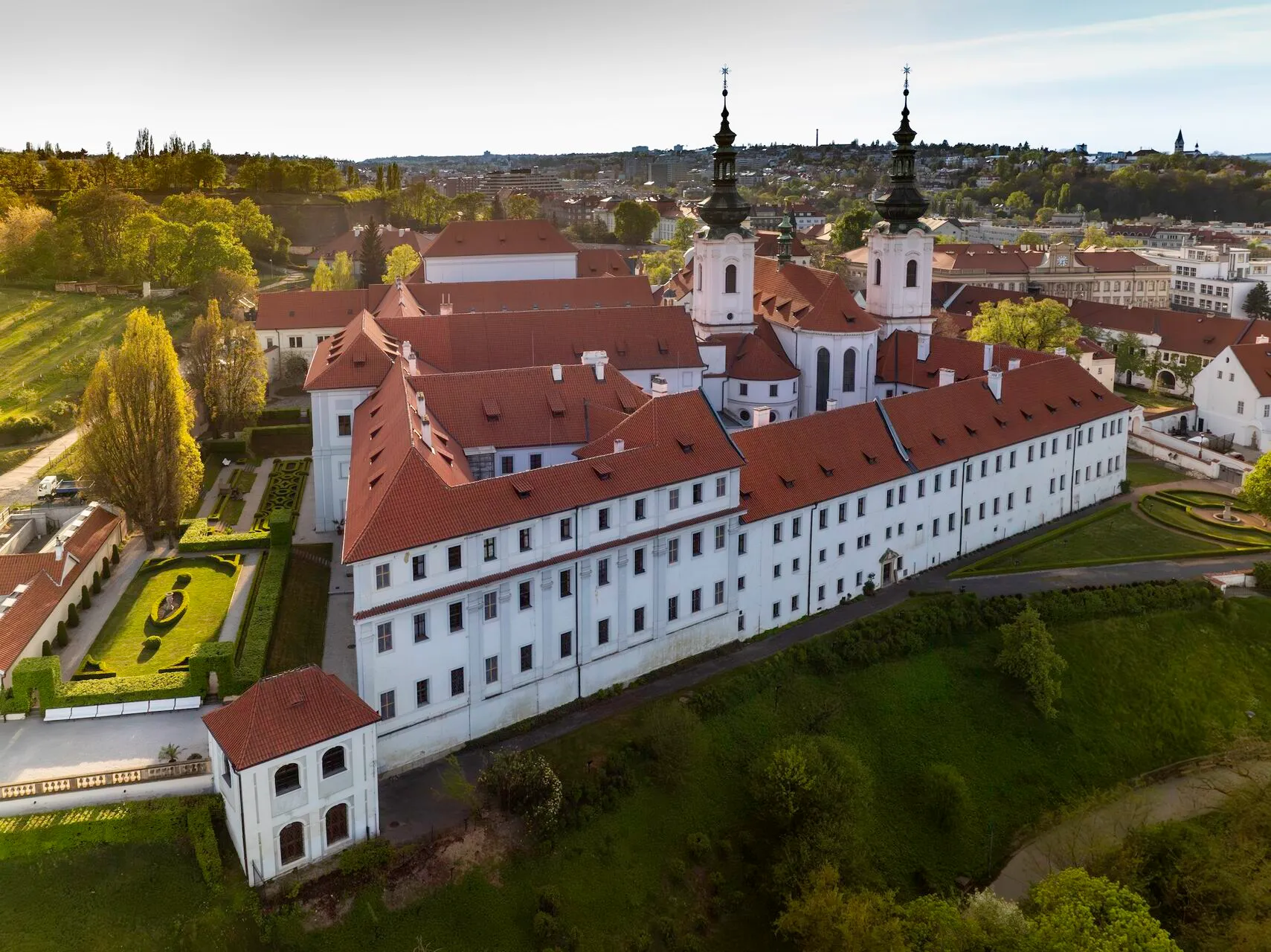 Widok z lotu ptaka na Klasztor na Strahowie z białymi budynkami, czerwonymi dachami i podwójnymi wieżami na wzgórzu.