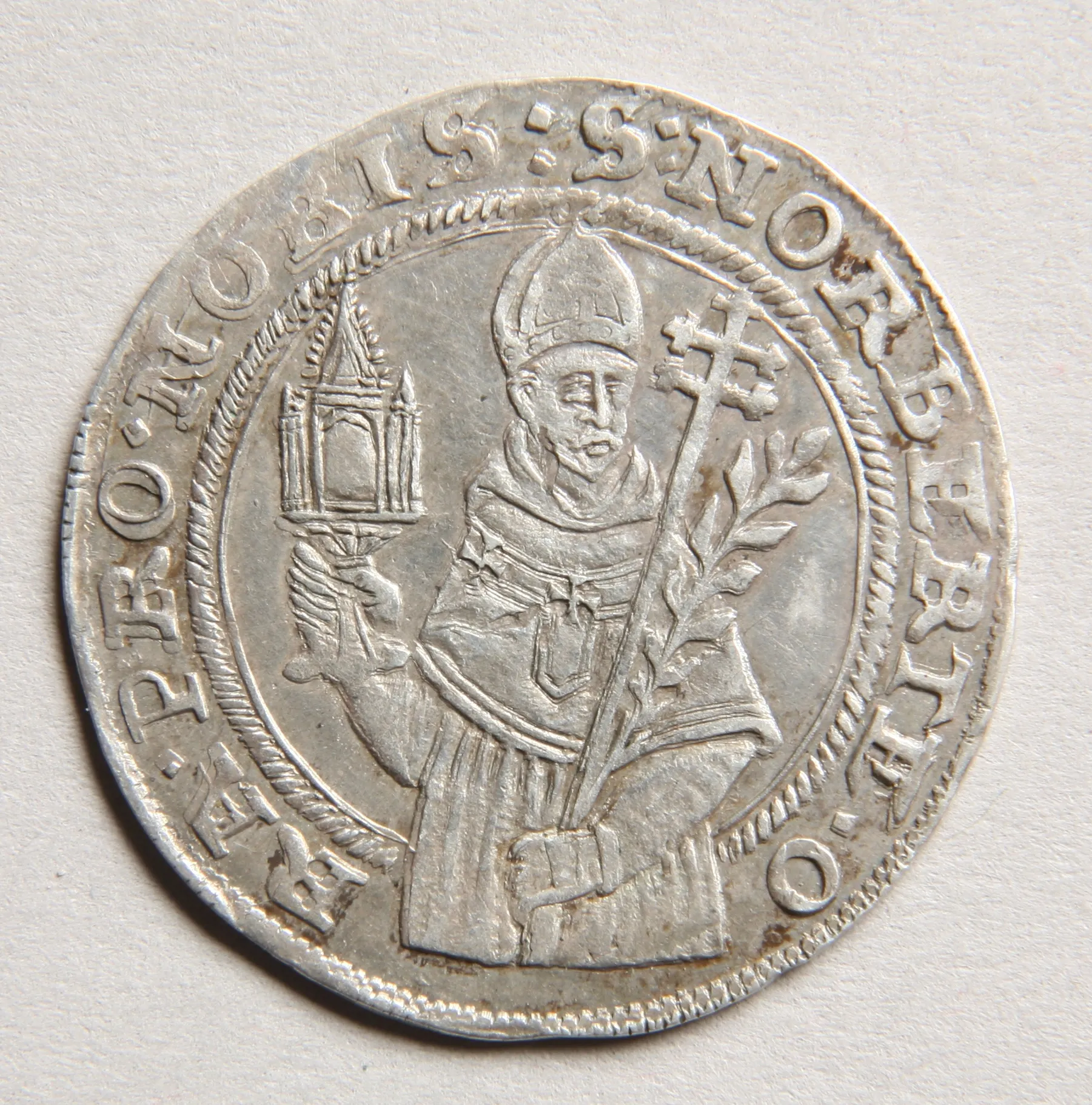 silver coin with a figure of a bishop holding a cross and a church model, surrounded by Latin text