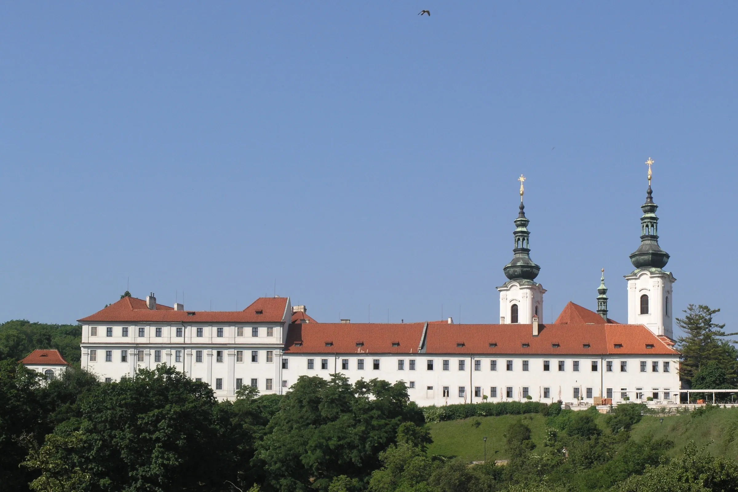 White monastery building with red roof and two green spires on a hillside under a clear blue sky