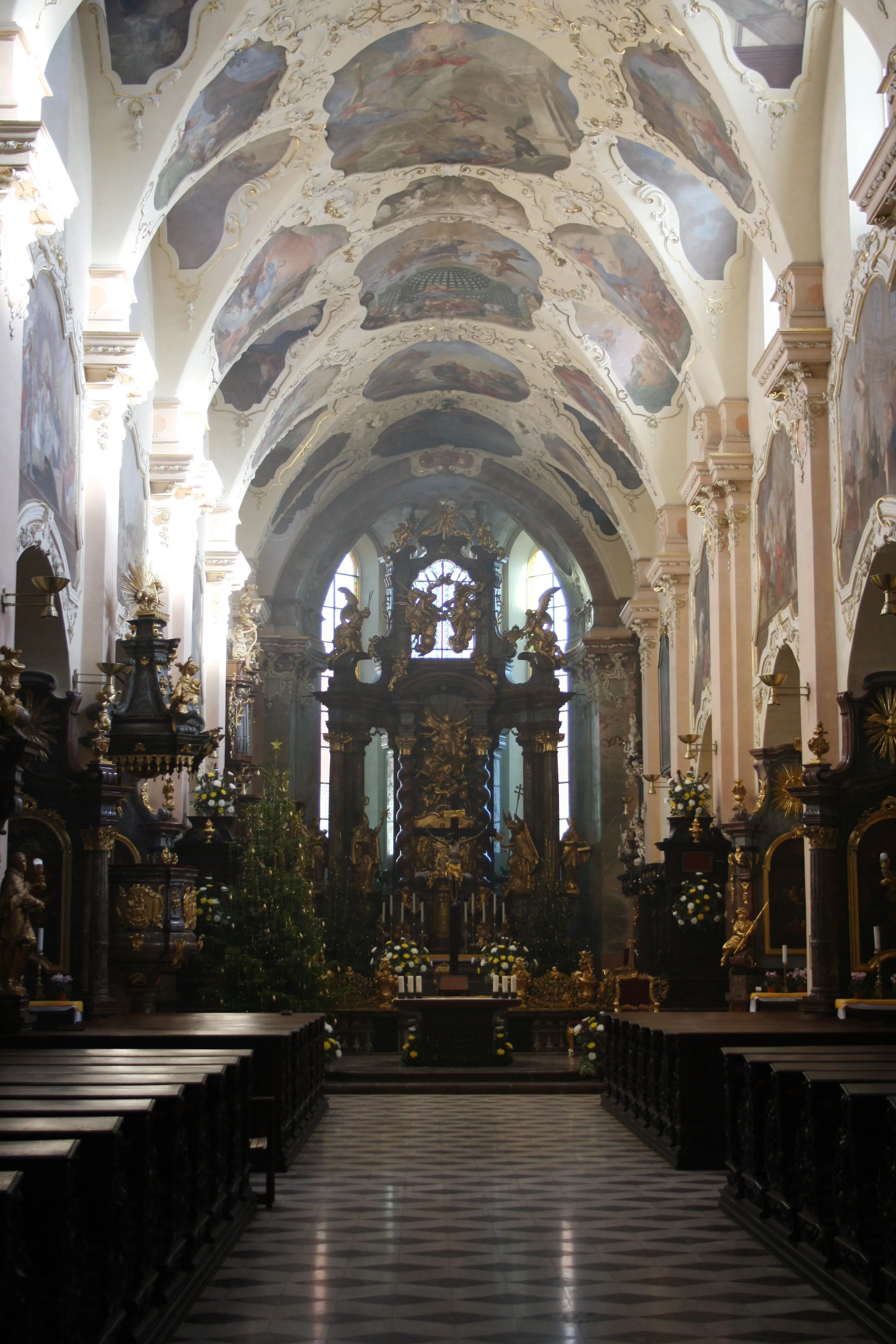 Interior of Strahov Monastery with a vaulted ceiling painted with frescoes, a central altar, and dark wooden pews.
