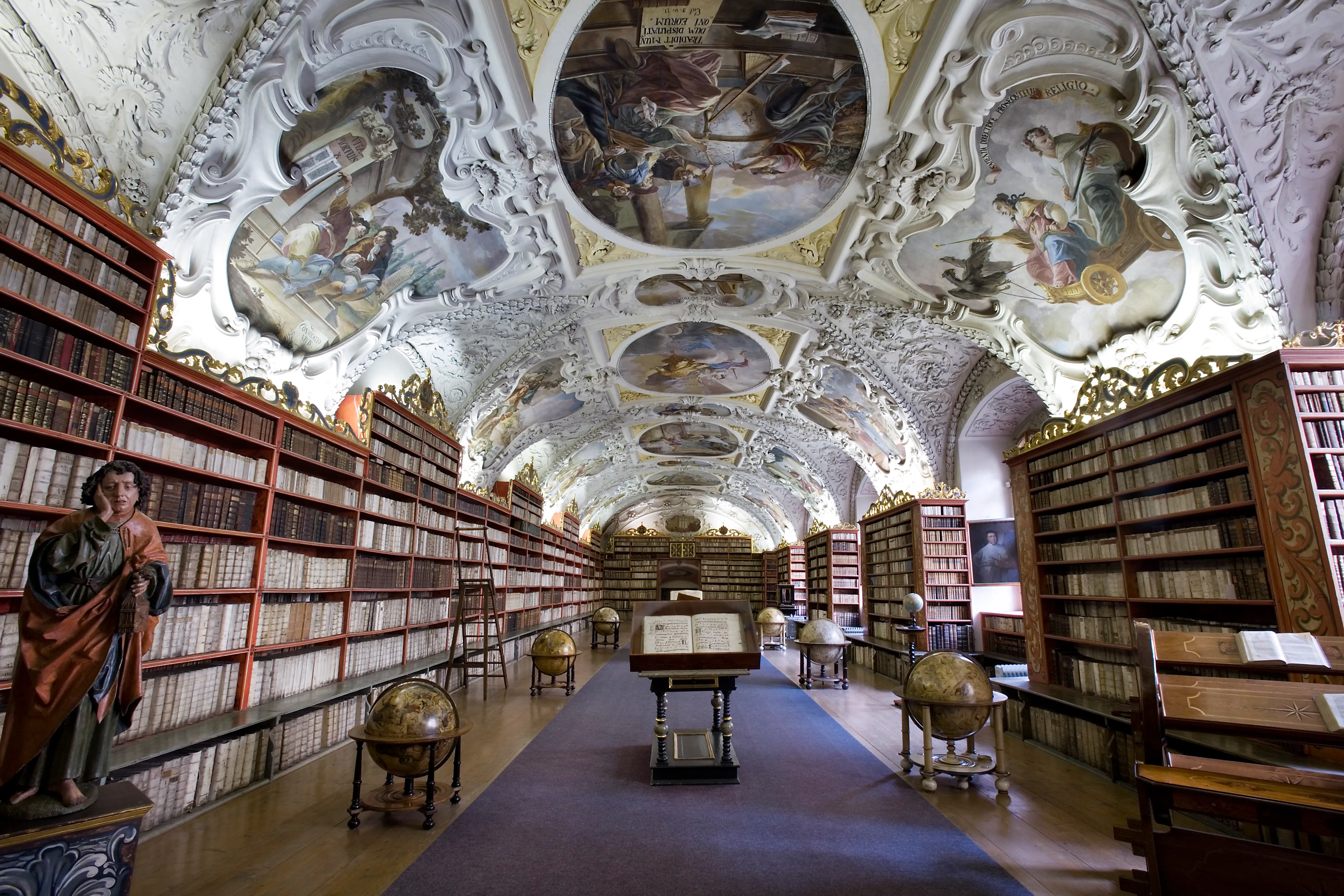 Baroque library hall with ornate painted ceiling frescoes, tall wooden bookshelves filled with books, globes, and a statue of a robed figure