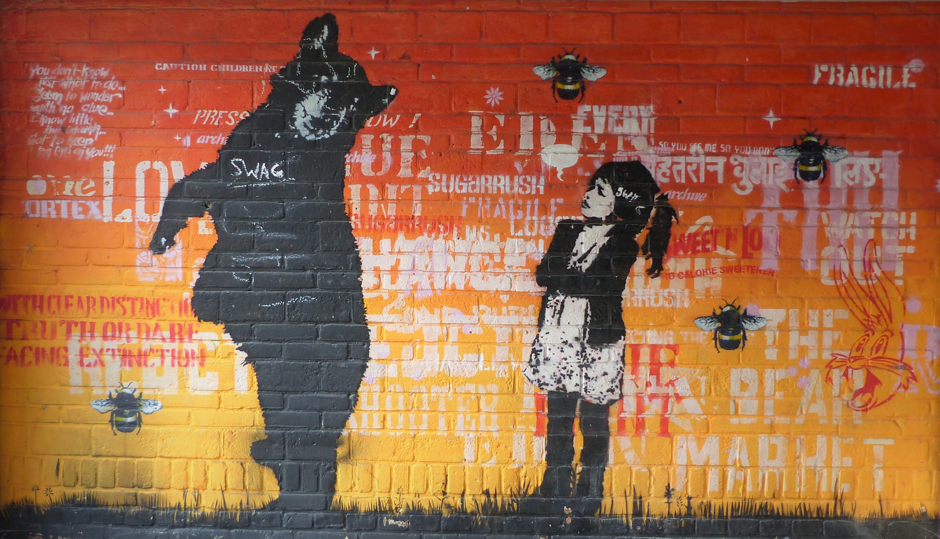 Vibrante mural de pared de ladrillos con una gran silueta de oso negro, una figura de niña, frases de texto multilingües y motivos de abejas en tonos rojos, amarillos y negros