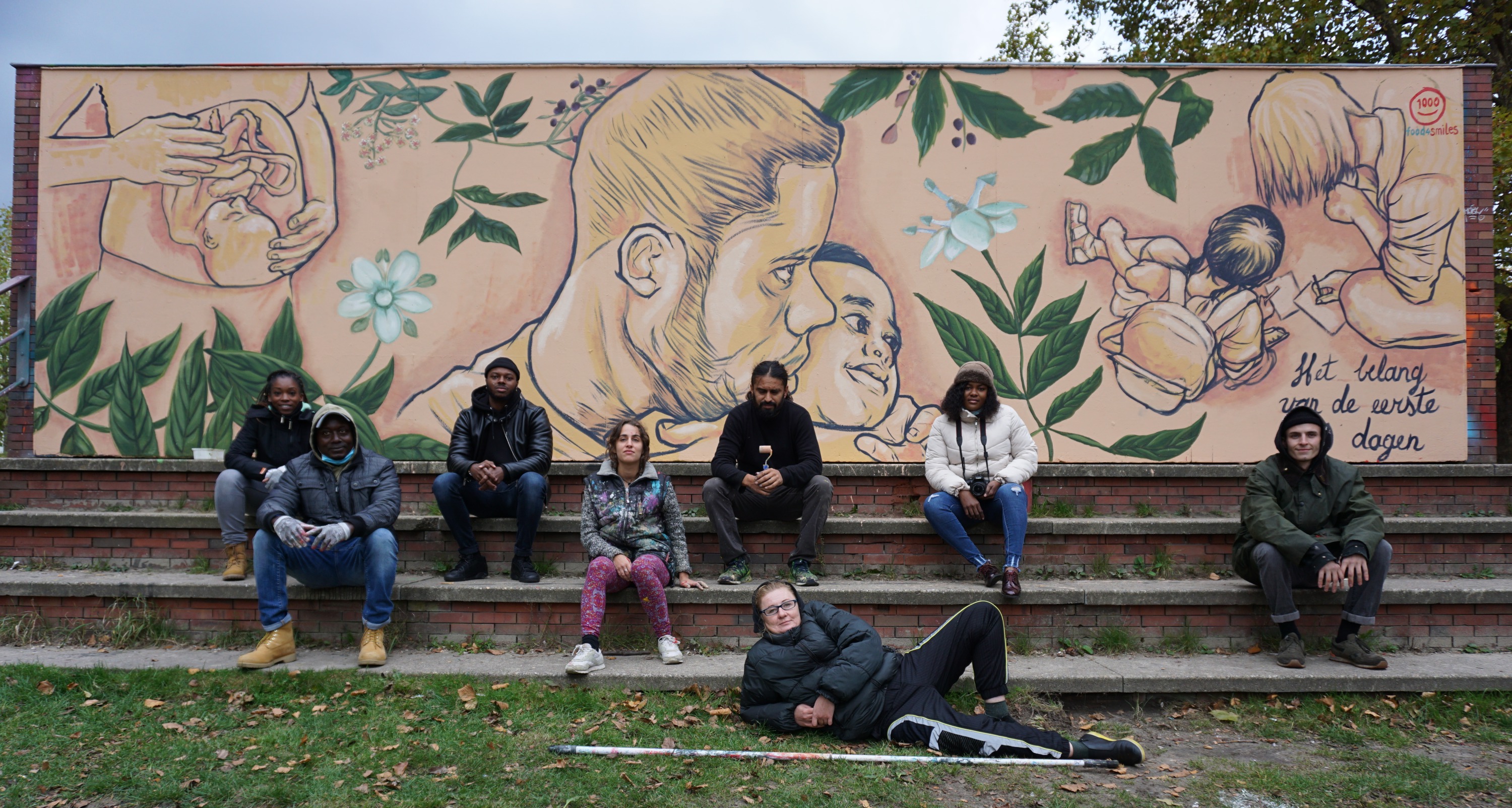Group of people seated on stone steps before a large mural featuring portraits, floral motifs, and Dutch text 'Mijn lichaam is een straat voor de toekomst'
