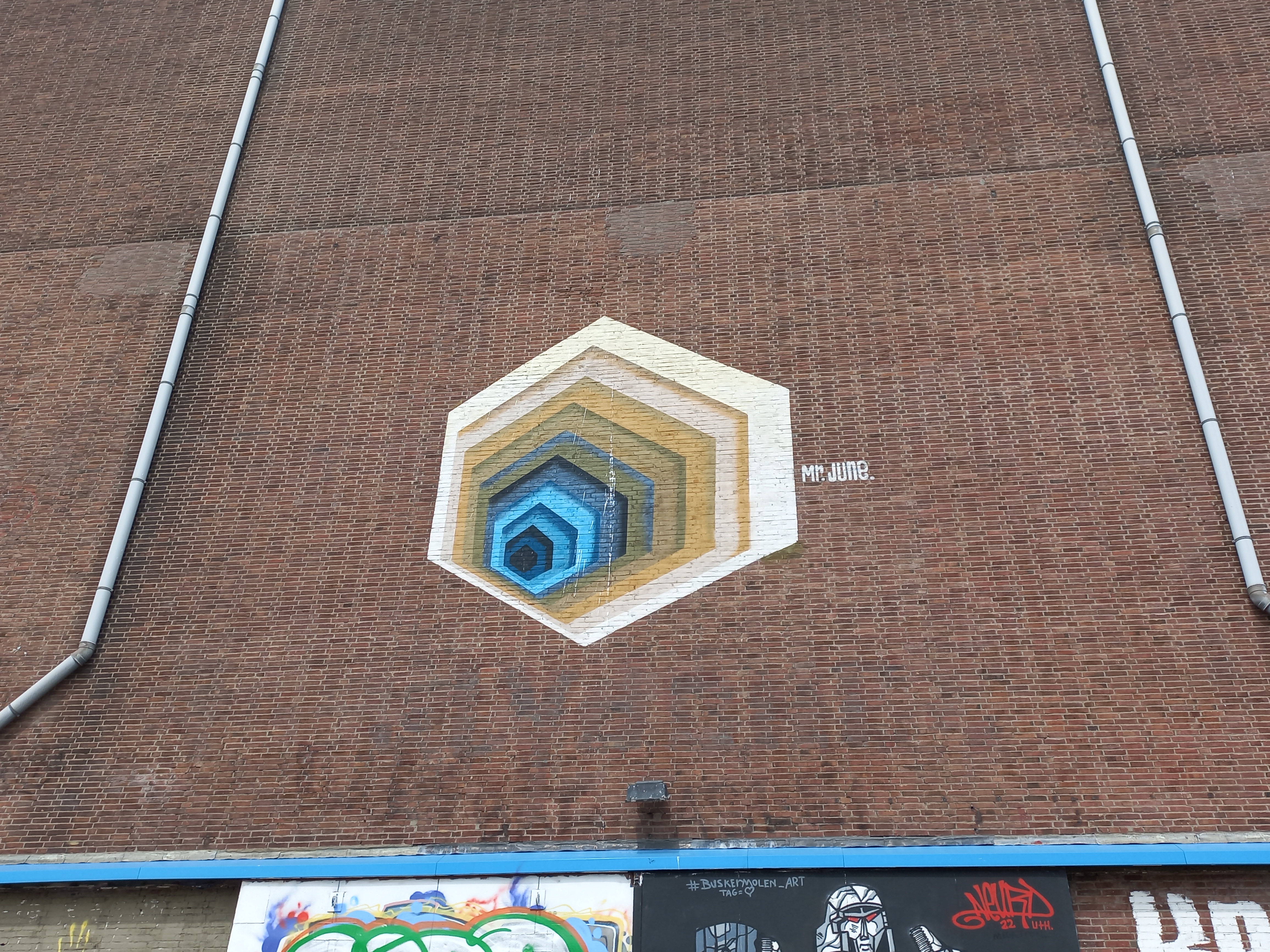 Mural hexagonal geométrico con formas concéntricas azules, amarillas y marrones pintado en una pared de ladrillos, con arte callejero adicional debajo