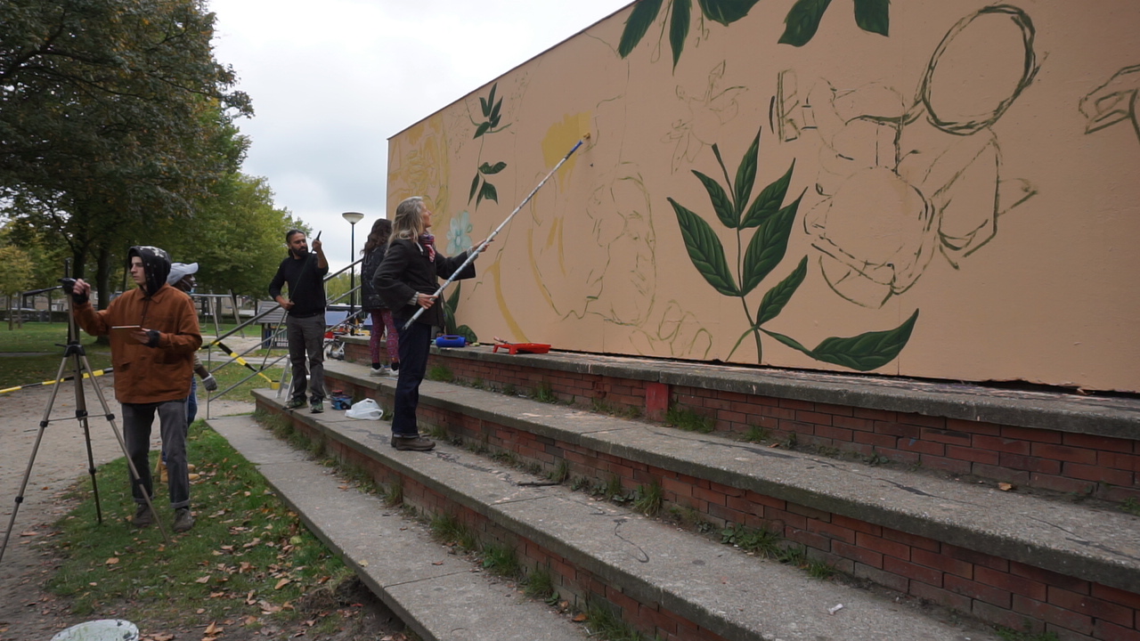 Artistas pintando un mural de arte callejero con motivos de plantas verdes en una pared beige, con gente trabajando en escalones de ladrillo y una zona de césped