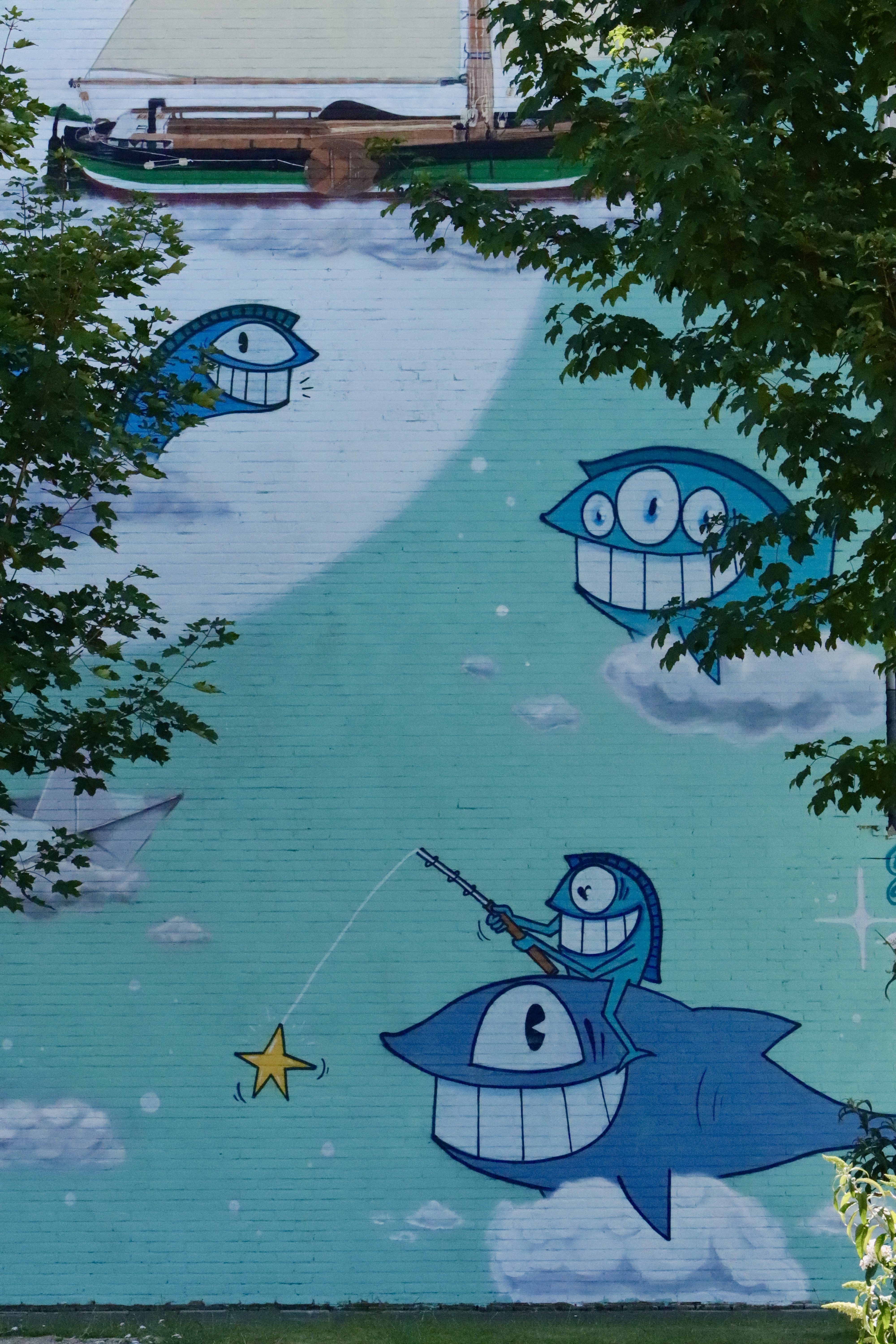 Mural azul vibrante sobre pared de ladrillo con personajes de peces de dibujos animados, uno pescando una estrella, con árboles enmarcando la obra.