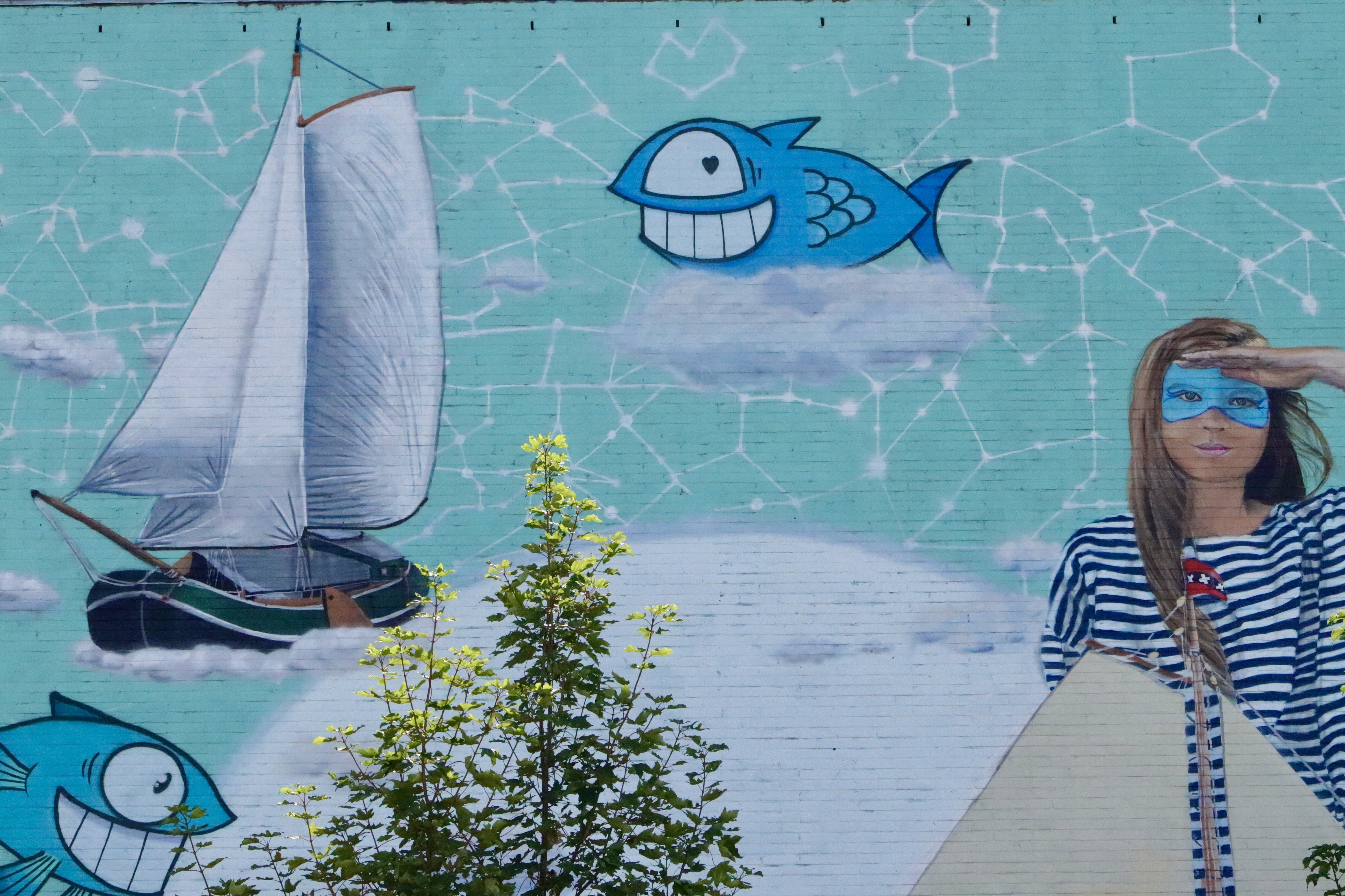 Vibrant street art mural featuring sailboat, smiling blue fish, constellation patterns, and masked figure in striped shirt against turquoise background