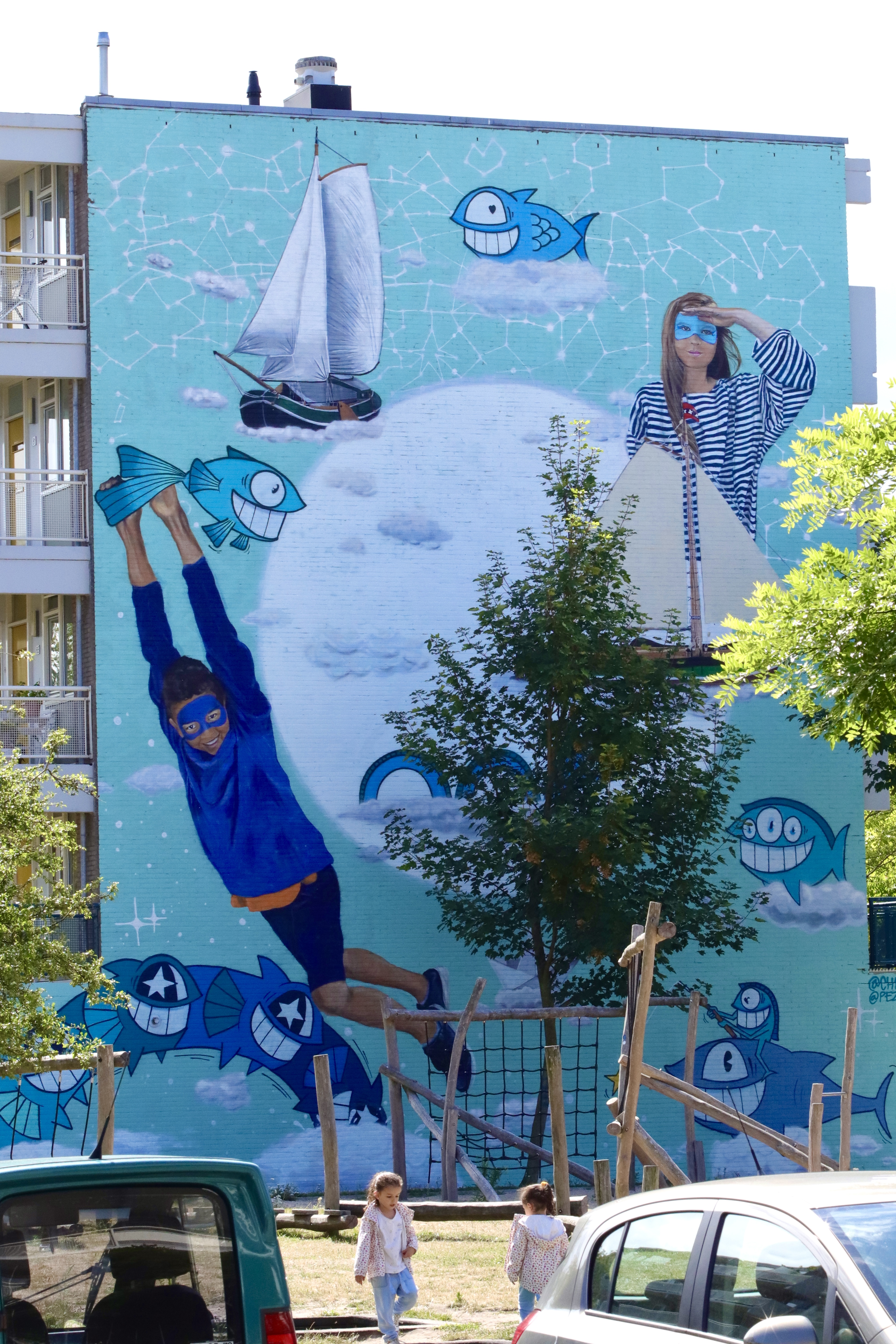 Vibrant turquoise mural featuring blue cartoon fish, a sailboat, moon, and people in blue outfits on a building exterior, with trees and cars below.