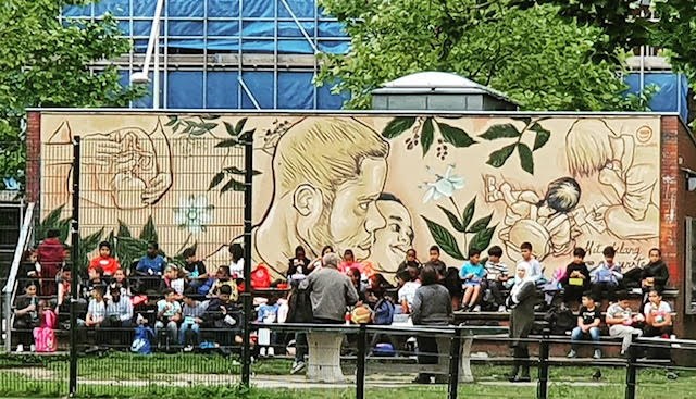 Gran mural al aire libre con figuras de línea y follaje, multitud reunida al frente observando un evento cerca de la obra de arte