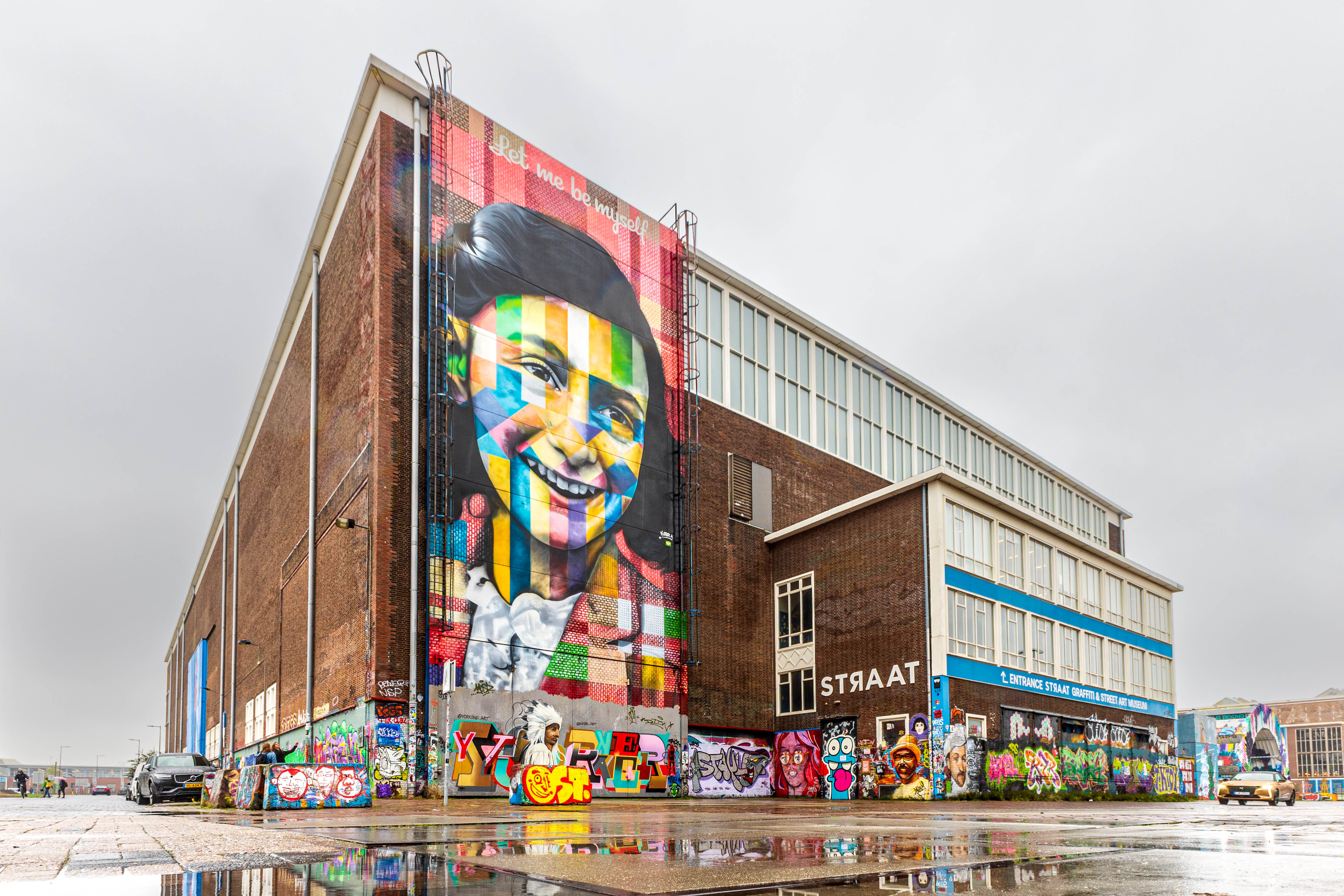 Gran mural colorido de una cara sonriente en el costado de un edificio de ladrillos con la palabra STRAAT en la entrada