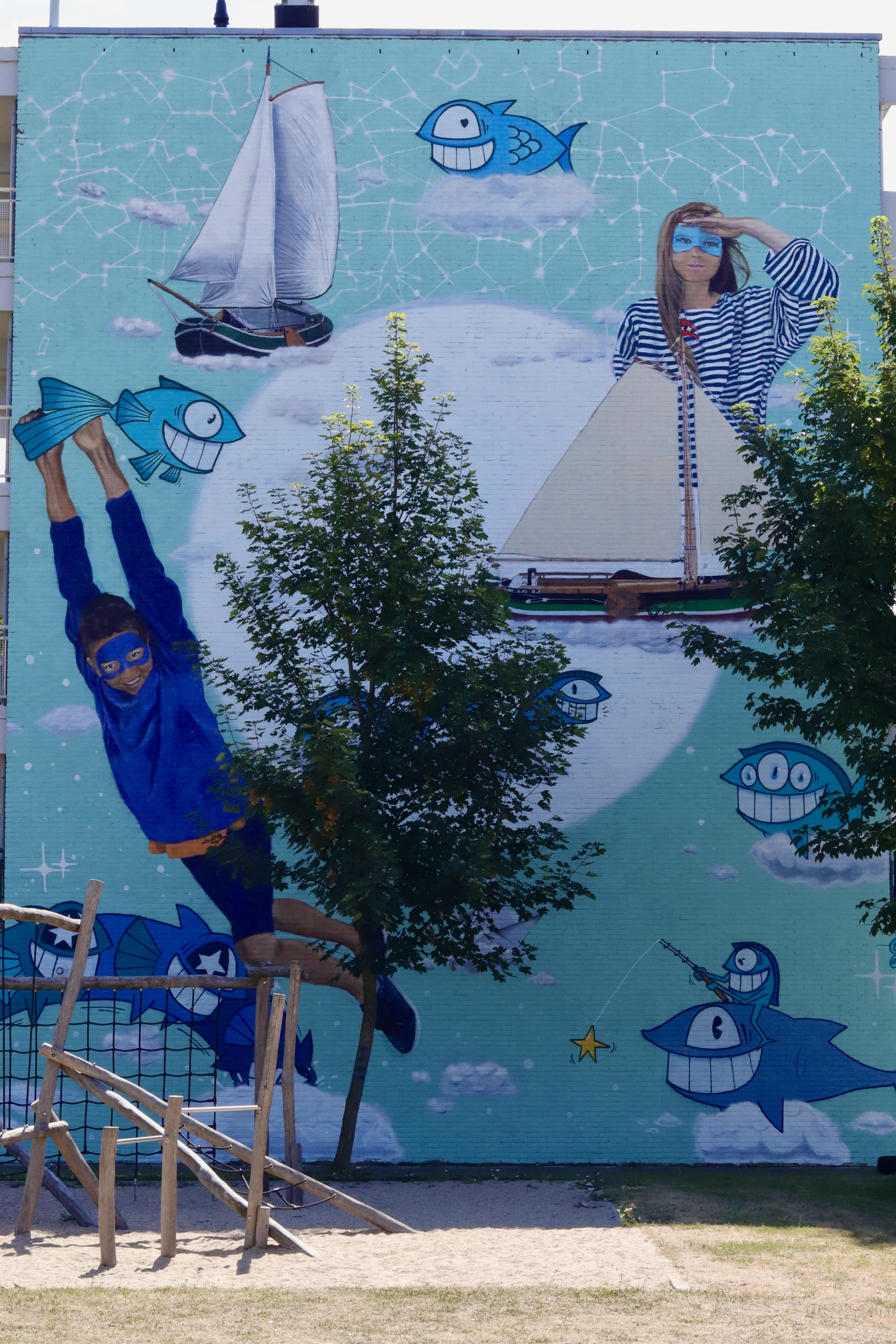 Vibrante mural turquesa con personajes de dibujos animados, peces azules sonrientes, veleros y patrones de constelaciones en una pared exterior.