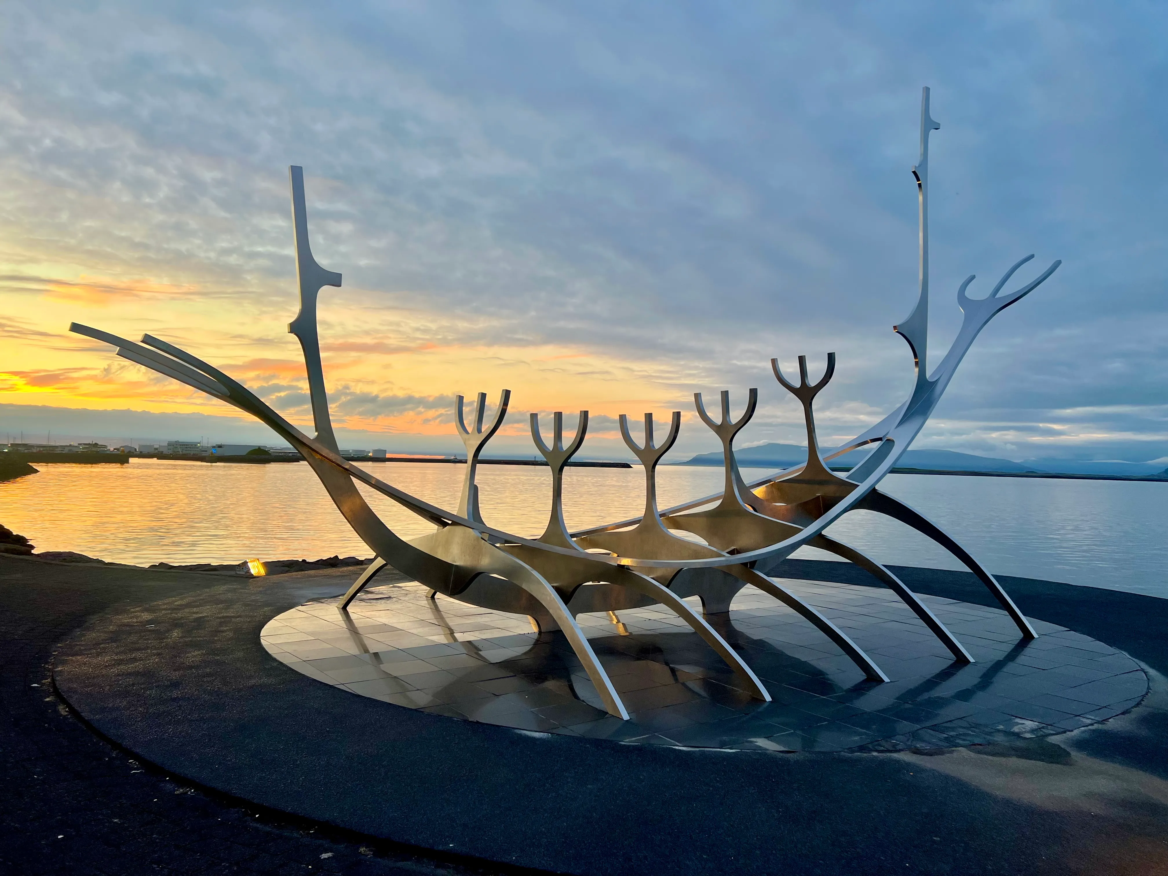 Rzeźba Sun Voyager ze stali nierdzewnej przypominająca statek wikingów na nabrzeżu Reykjavíku o zachodzie słońca, ze złotym niebem odbijającym się w spokojnych wodach portu