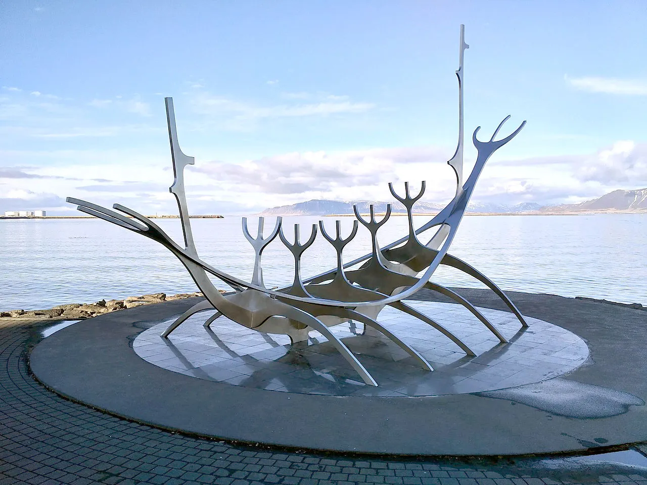 Rzeźba Sun Voyager ze stali nierdzewnej na okrągłej betonowej platformie na nabrzeżu Reykjavíku, ze spokojnym morzem i odległymi górami pod błękitnym niebem