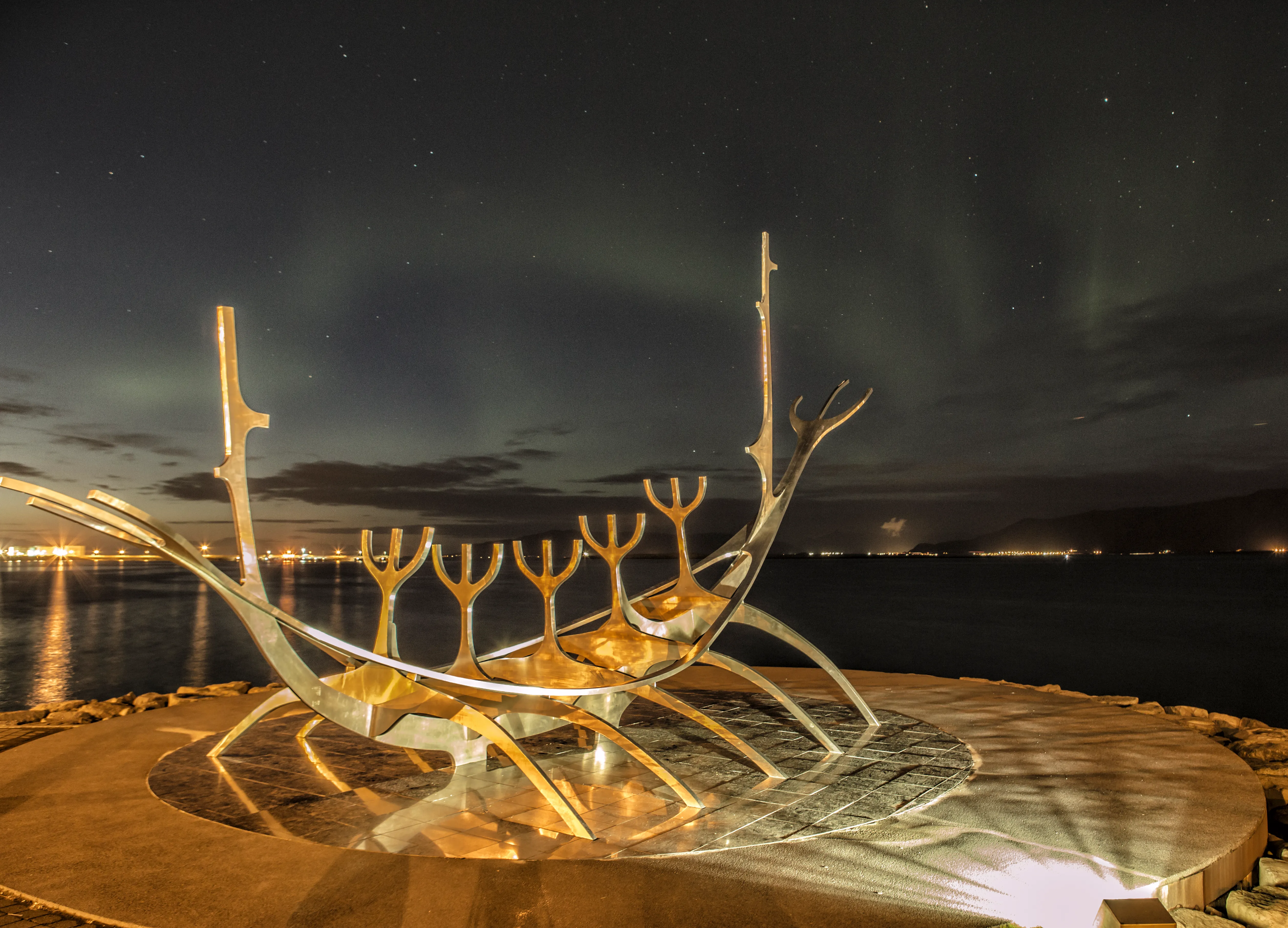 Nocny widok na podświetloną rzeźbę ze stali nierdzewnej Sun Voyager na nabrzeżu w Reykjavíku, z zorzą polarną świecącą na zielono na niebie.