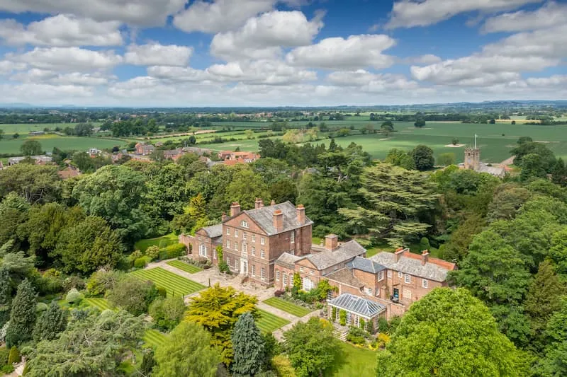 Vista aérea de Sutton Park, una casa de campo georgiana de ladrillo rojo rodeada de árboles maduros y jardines formales, con el campo y la aguja de una iglesia visibles a lo lejos bajo un cielo azul con nubes blancas.
