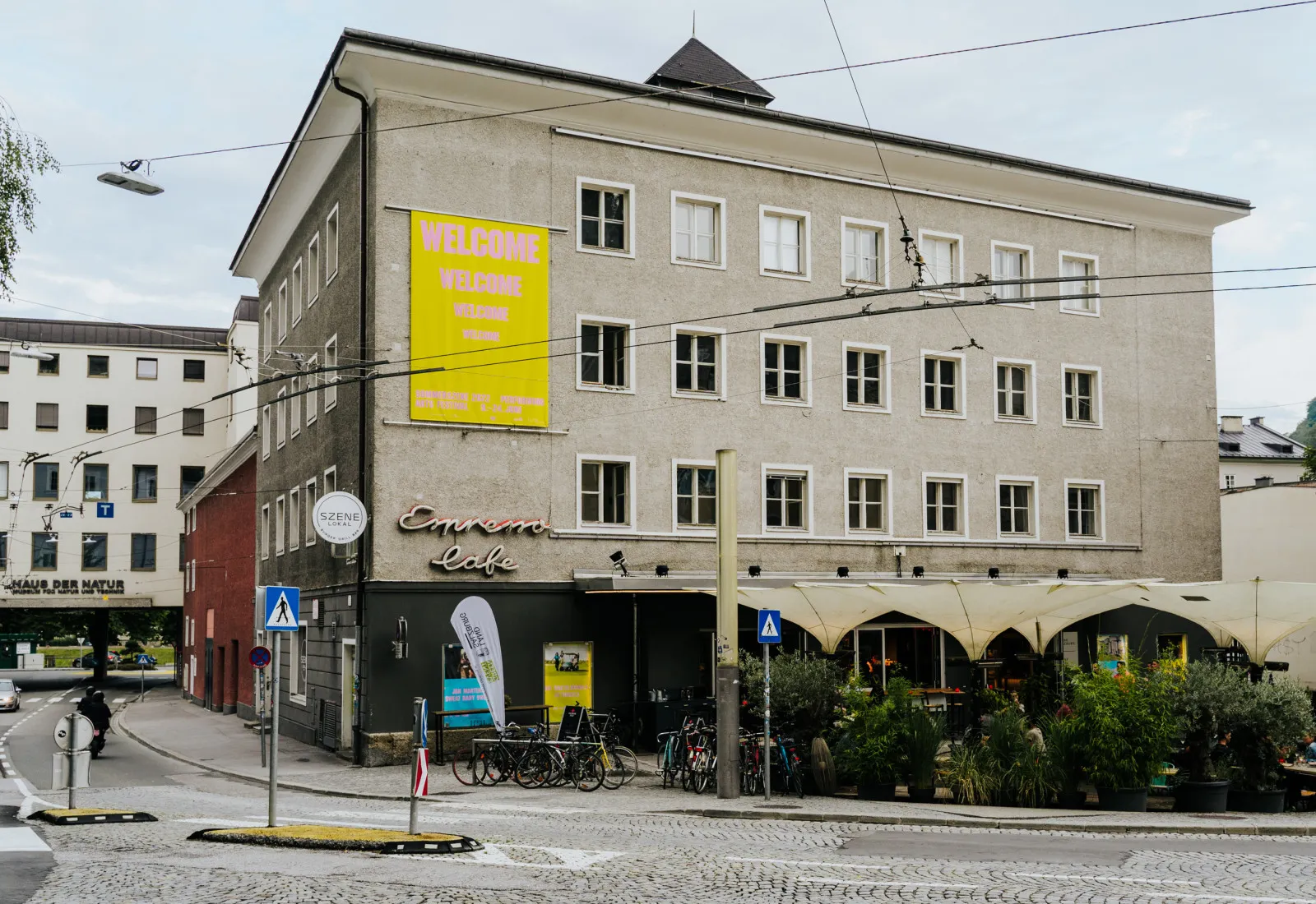 Widok ulicy na wielopiętrowy, szary budynek z dużym żółtym banerem 'Welcome to Szene Salzburg' oraz szyldem Cinema Café na parterze, z rowerami zaparkowanymi na zewnątrz i przewodami trakcji tramwajowej nad głową.