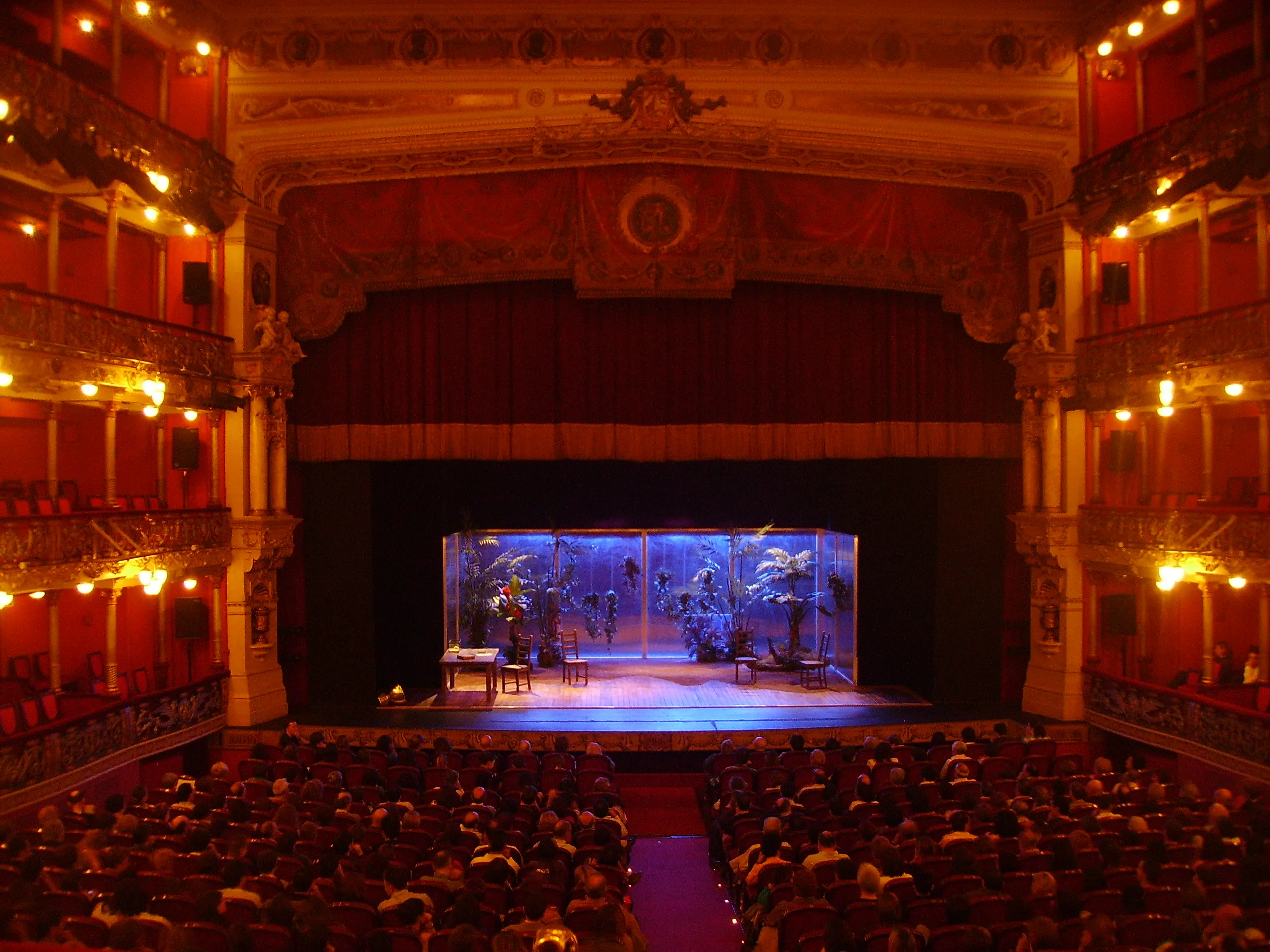 Vista del auditorio del Teatro Arriaga con el aforo completo durante una función en un escenario iluminado de azul, enmarcado por balcones ornamentados y una cálida iluminación.