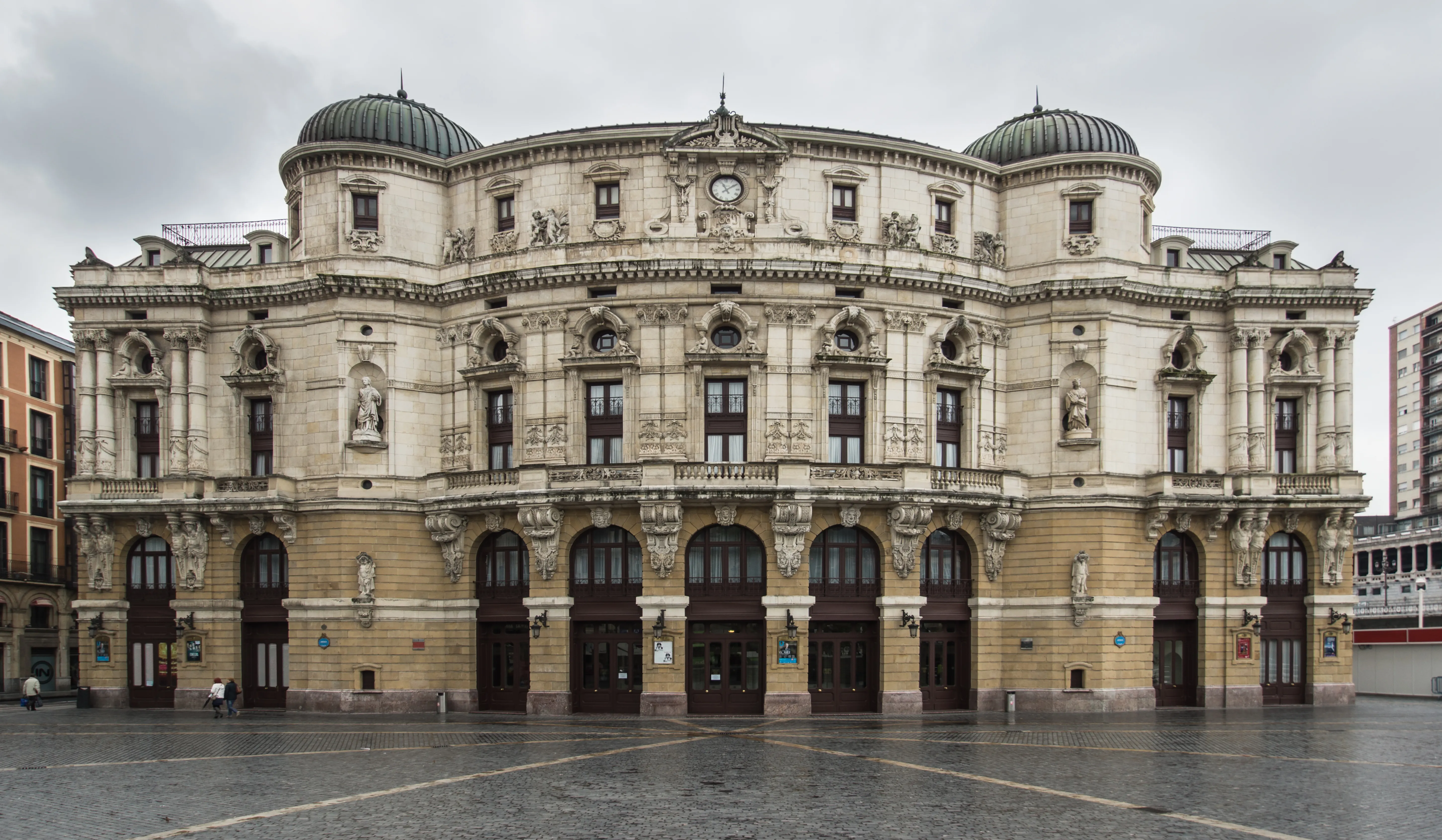 Vista exterior amplia del Teatro Arriaga Antzokia, un gran teatro neobarroco con dos torres en esquina rematadas en cúpulas, entradas arqueadas y una ornamentada fachada de piedra frente a una plaza pavimentada.