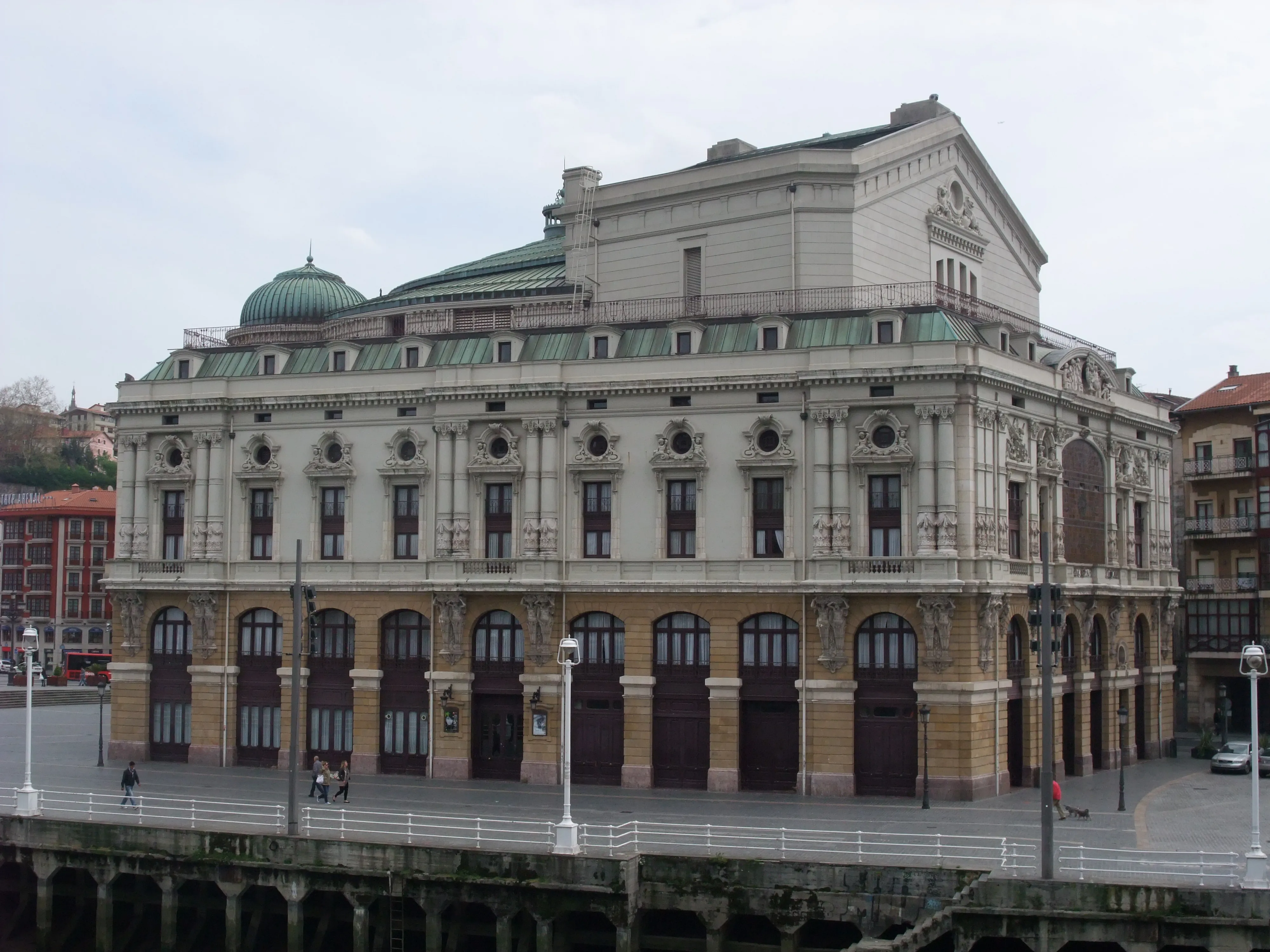 Vista exterior del Teatro Arriaga, un gran edificio neobarroco con cúpula verde, ventanas arqueadas en la planta baja y una fachada ornamentada a lo largo de un paseo junto a la ría en Bilbao
