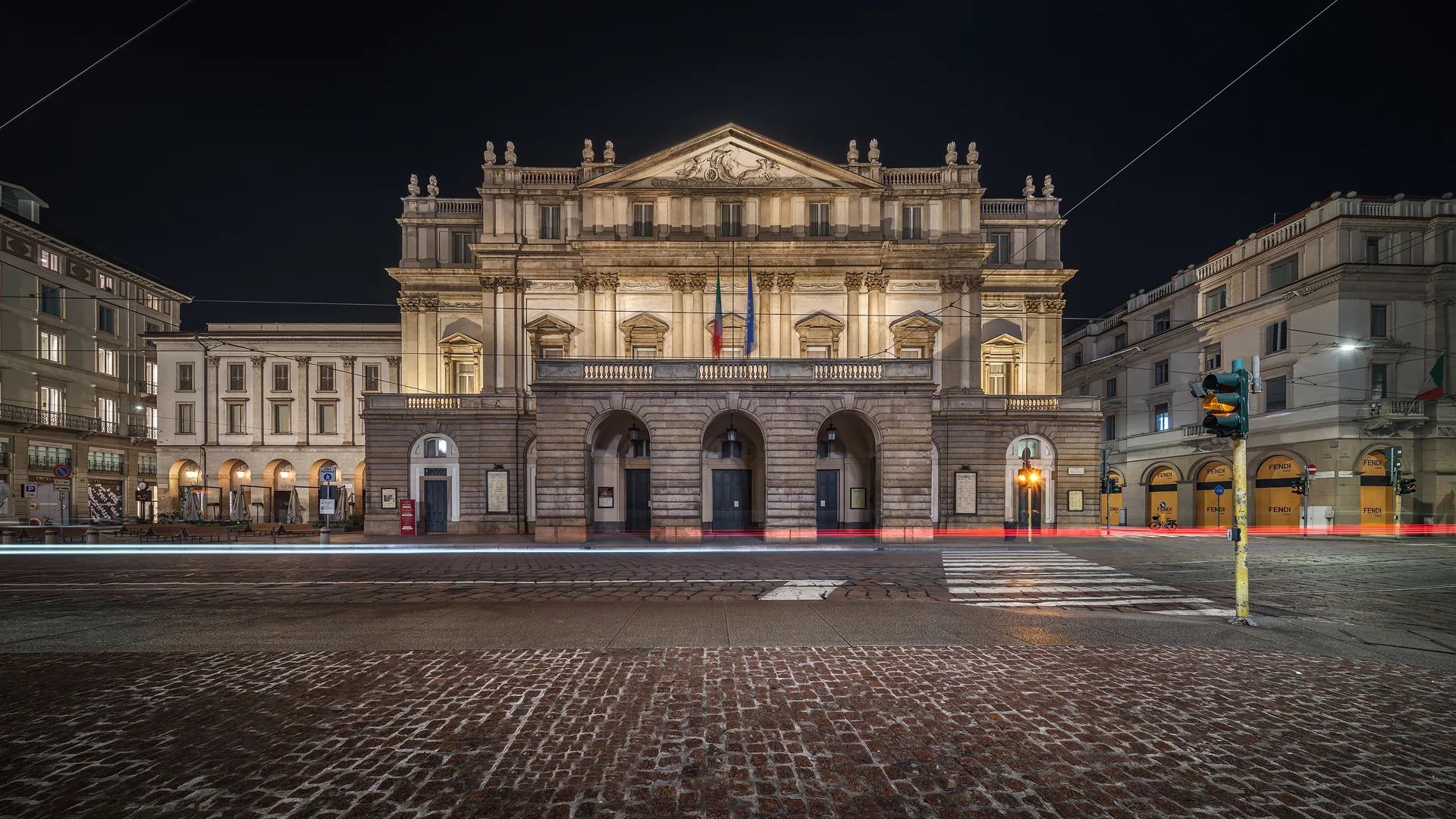 Gran fachada de teatro neobarroco iluminada de noche con entradas arqueadas, columnas y un frontón triangular, vista desde una plaza empedrada con estelas de luz de los vehículos que pasan
