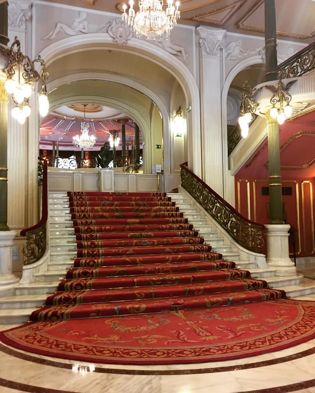 Gran escalinata de mármol con alfombra roja estampada, barandillas de hierro ornamentadas y lámparas de araña de cristal dentro del Teatro Arriaga