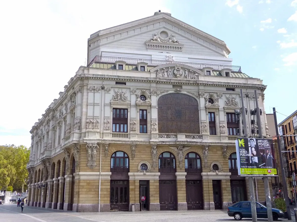 Grandioso edificio teatral de estilo neobarroco con una ornamentada fachada de color crema, ventanas arqueadas, mampostería decorativa y un gran frontón triangular, con una calle y un coche aparcado en frente.